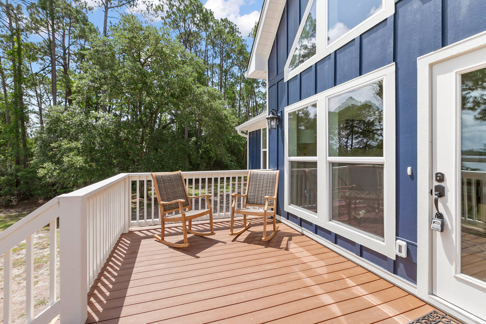 Wooden deck with two rocking chairs, white handrail, blue house siding, large windows, and surrounding trees