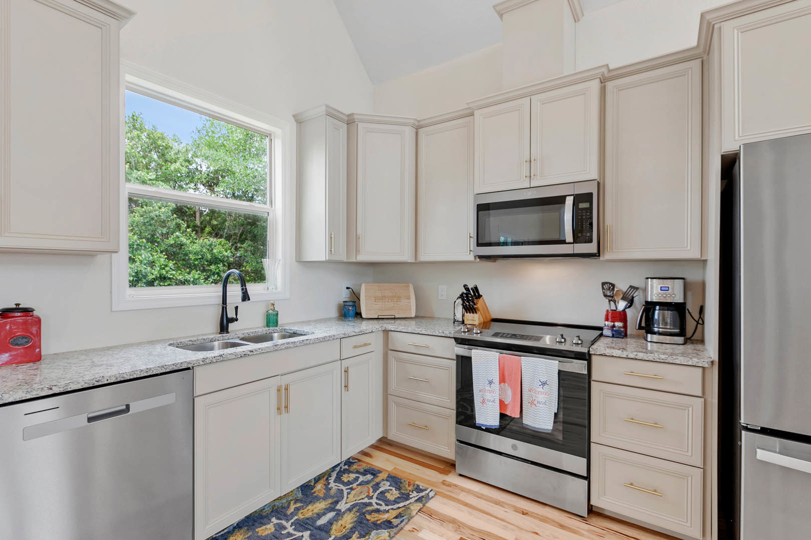 White kitchen cabinets, black faucet, built-in microwave, oven with hanging towel, window overlooking trees, white walls, light countertops