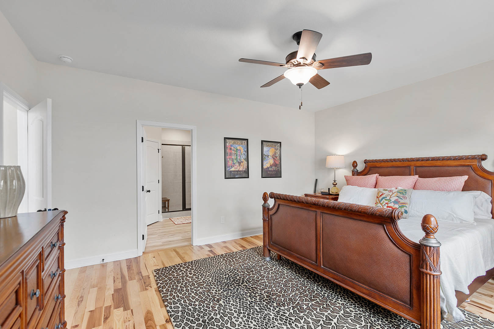 Bedroom with upholstered bed, white pillows, bedside lamp, wooden dresser, ceiling fan with light, framed street photograph, and framed painting on neutral walls.