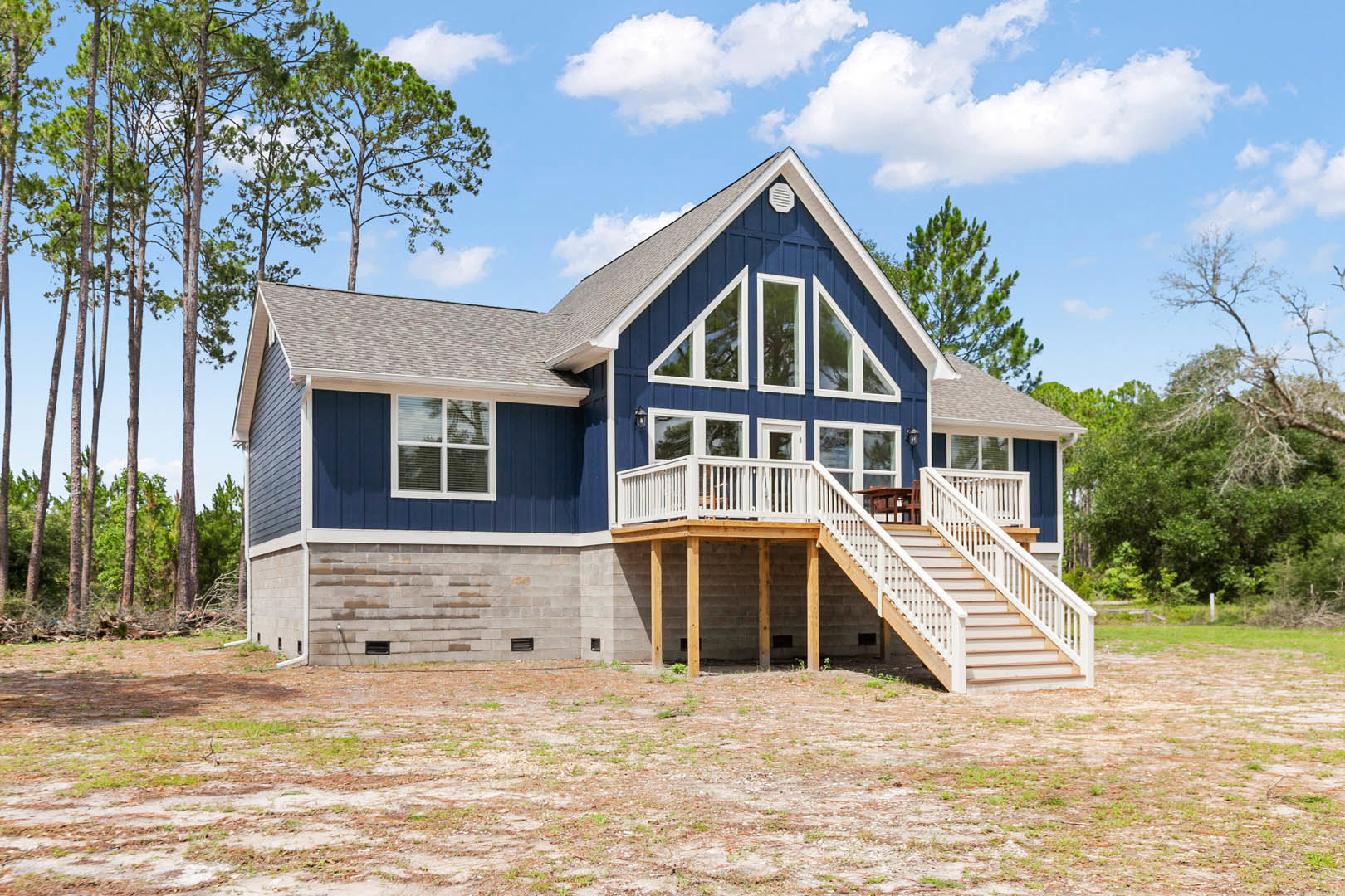 Blue siding house with white trim, elevated wooden deck, covered front porch, large windows, gabled roof, surrounded by trees and landscaping under a partly cloudy sky
