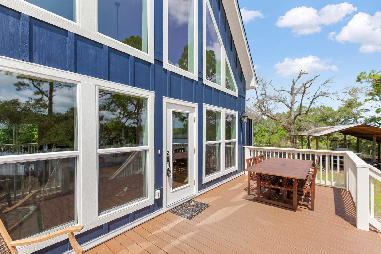Wood deck with black mat, outdoor table and chairs, blue house with white trim, large windows, surrounded by trees under partly cloudy sky