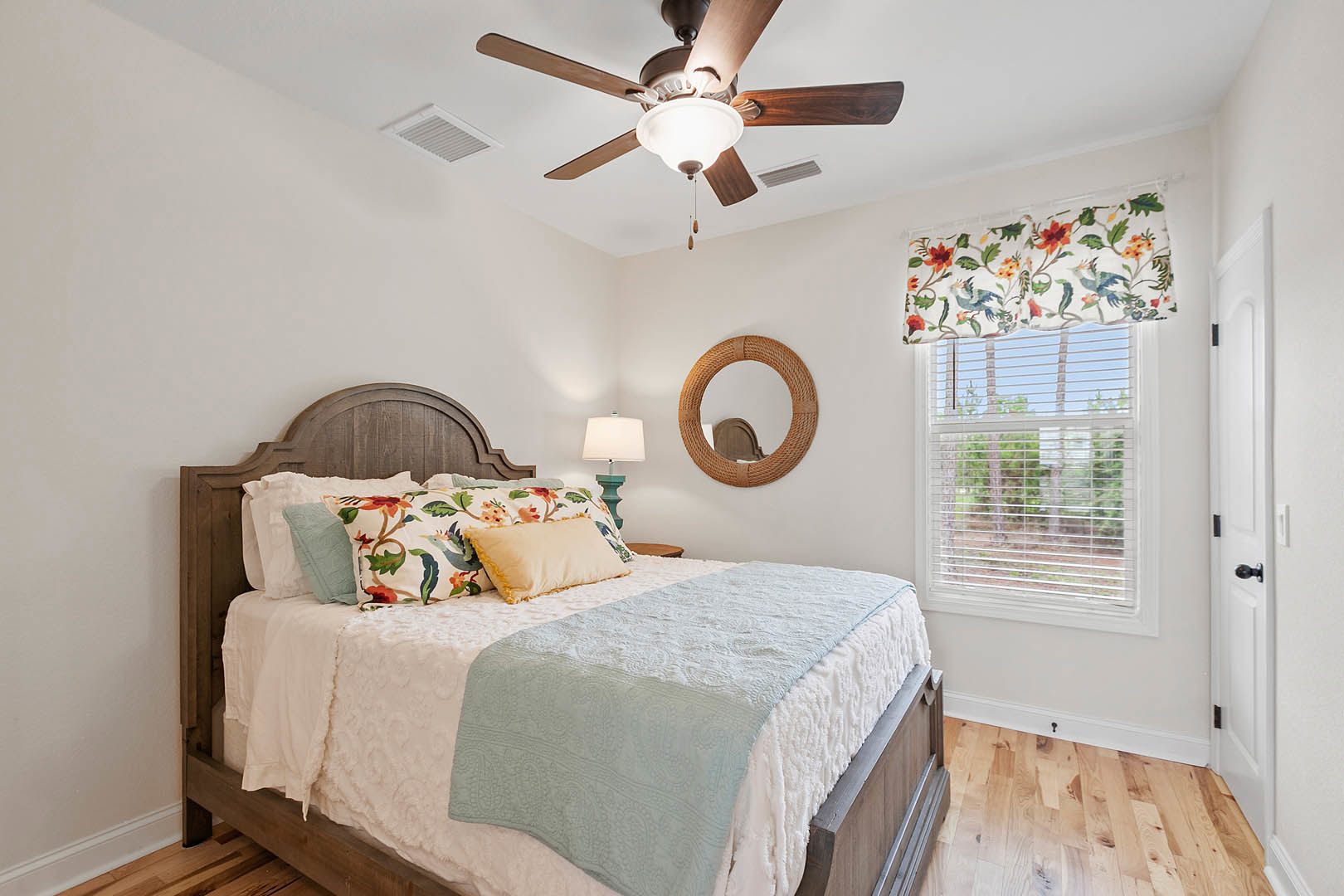 Bedroom with ceiling fan, upholstered bed, floral and bird patterned curtains, circular wall mirror, bedside lamp, and decorative pillows