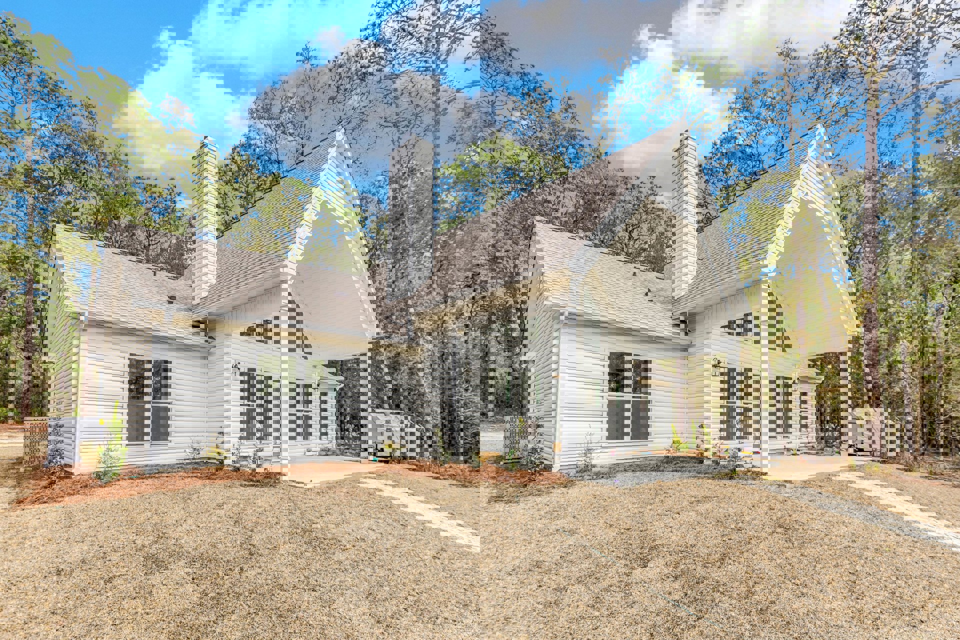 White two-story house with wide covered porch, grey metal gate, manicured lawn, brick chimney, screened windows, surrounded by trees under partly cloudy sky