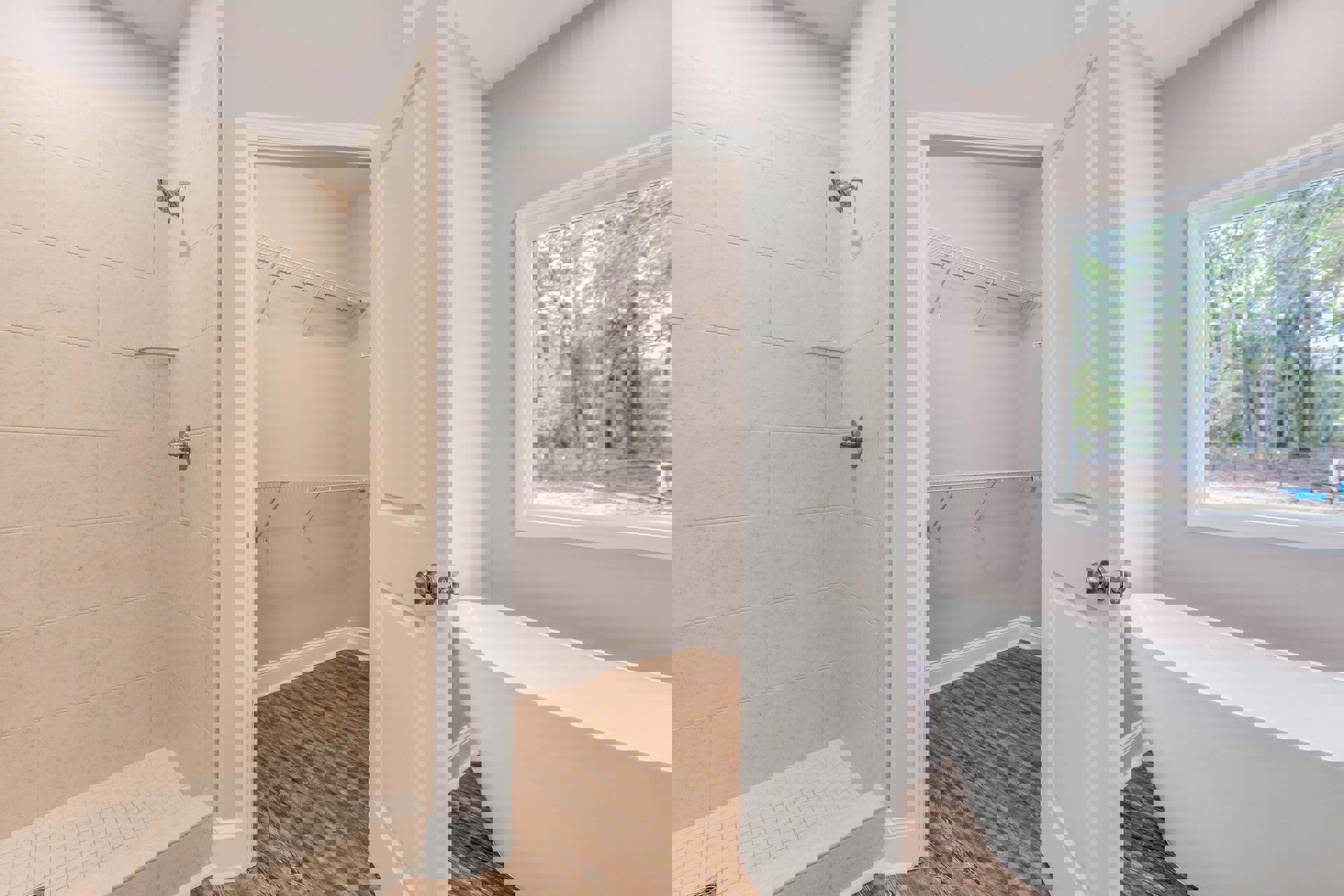 Bathroom with freestanding white tub, glass-enclosed shower featuring built-in shelf, large window overlooking trees, wood flooring, and modern door hardware