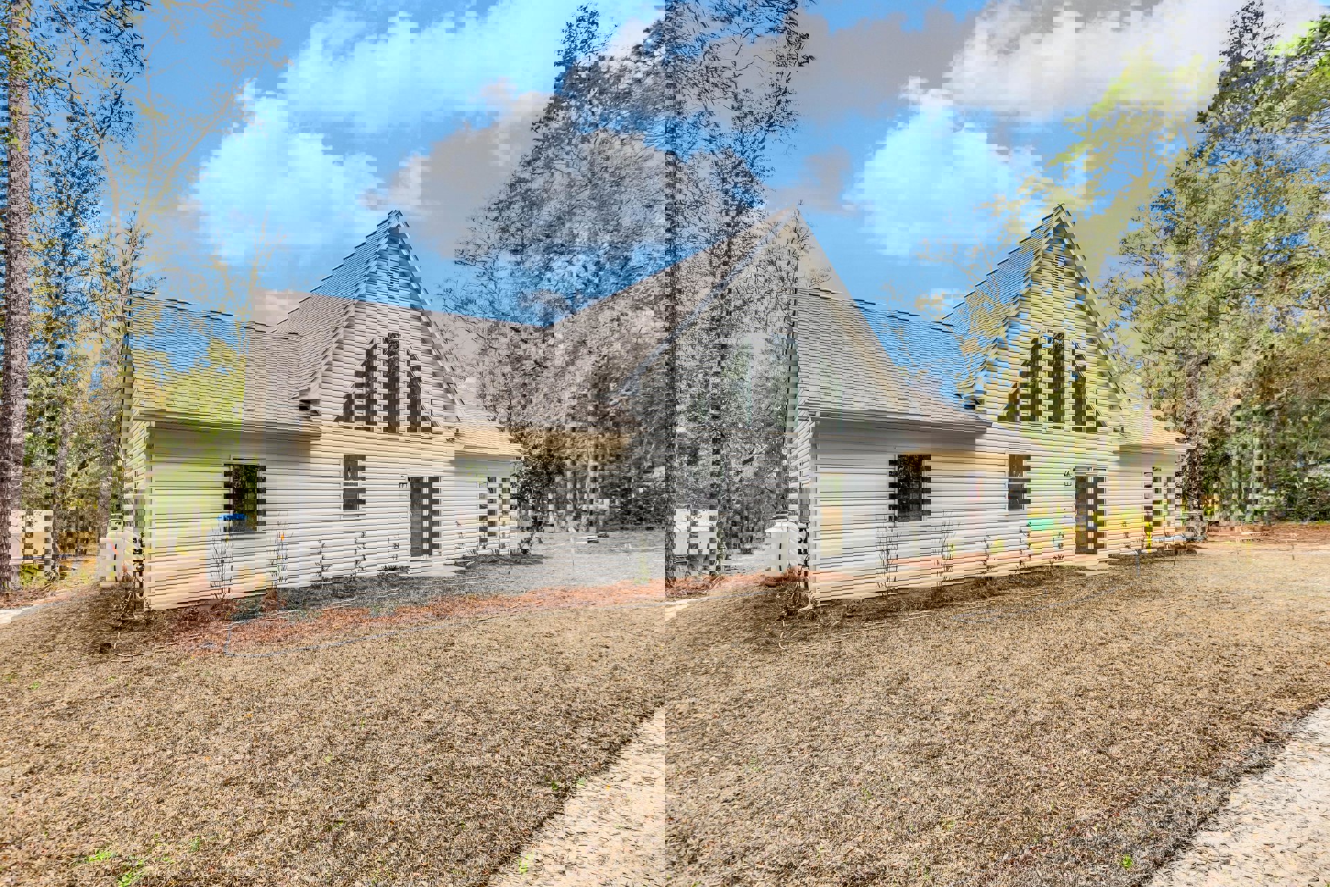 Two-story house with gray roof, white-framed windows, paved driveway, green lawn, and mature trees under a blue sky