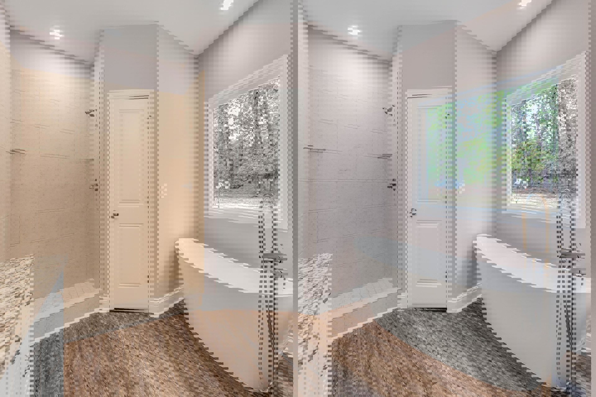 Bathroom with marble countertop, built-in bathtub and shower, white door with silver knob, window above sink overlooking trees, light tile flooring and walls.