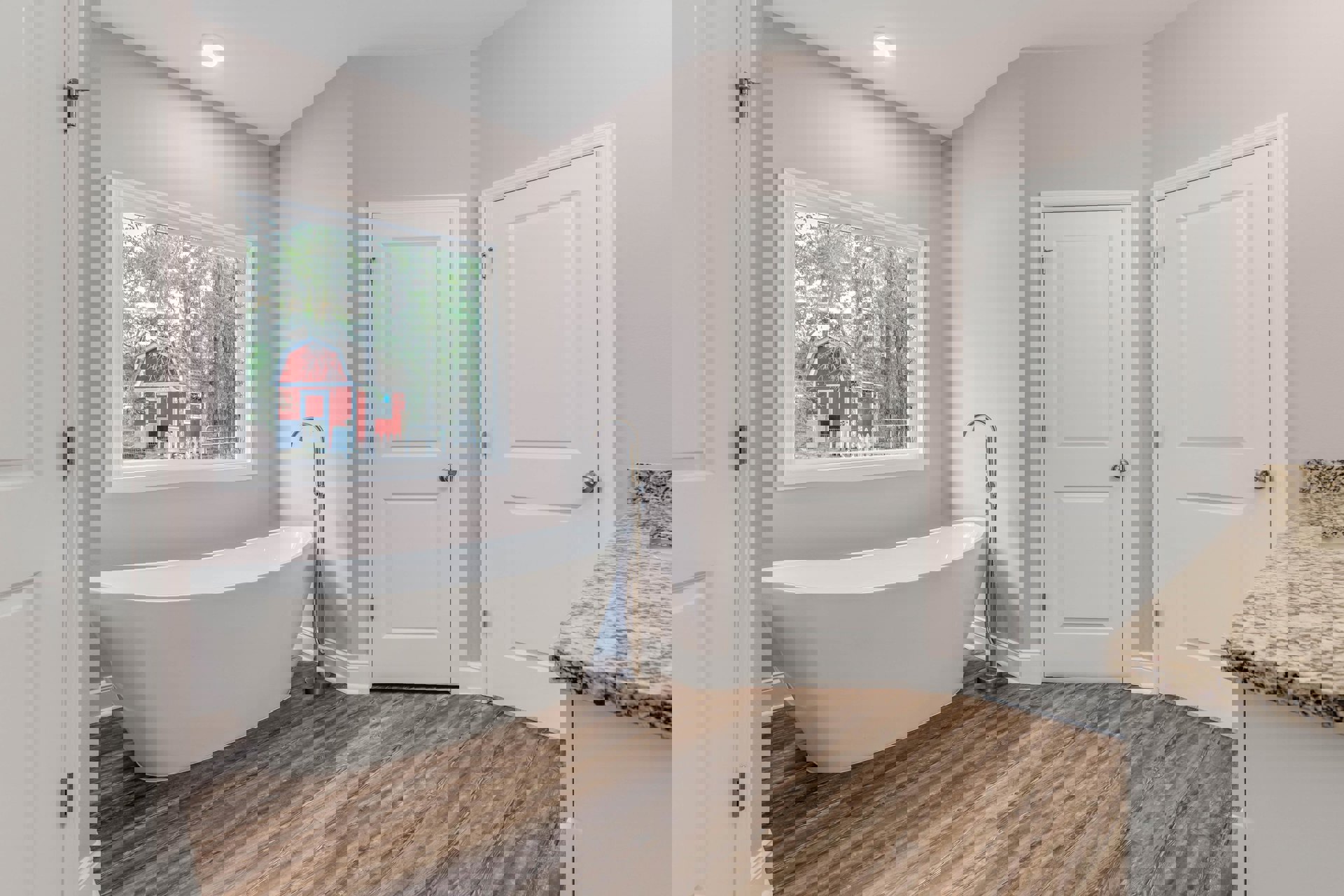 Freestanding white bathtub on wood floor beside white door with silver knob, window view of red barn, tile walls, chrome fixtures, and lamp.