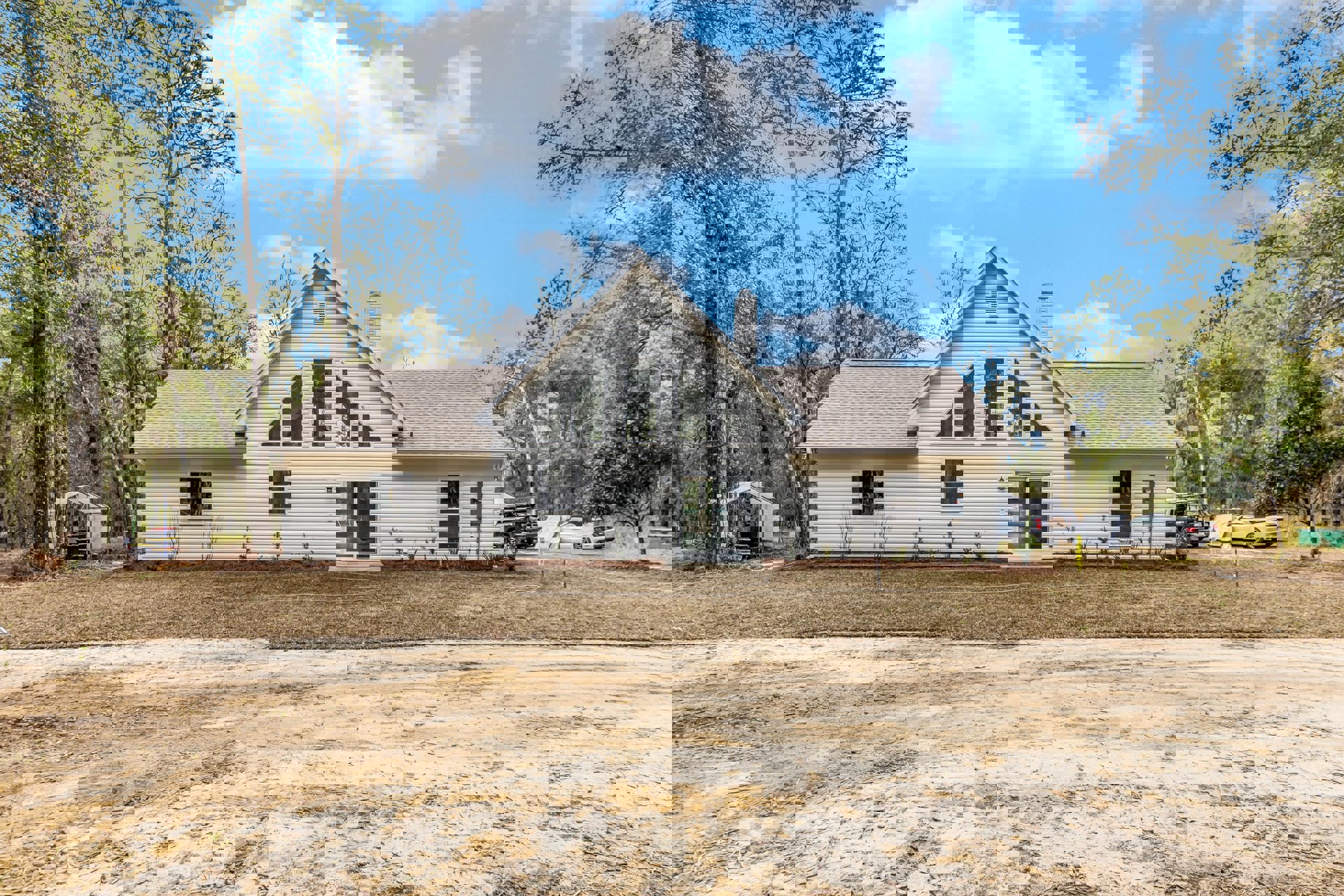 Modern cottage-style home with sliding glass door, large windows, and pitched roof, surrounded by trees and plants, white vehicle covered in plastic parked near dirt road under