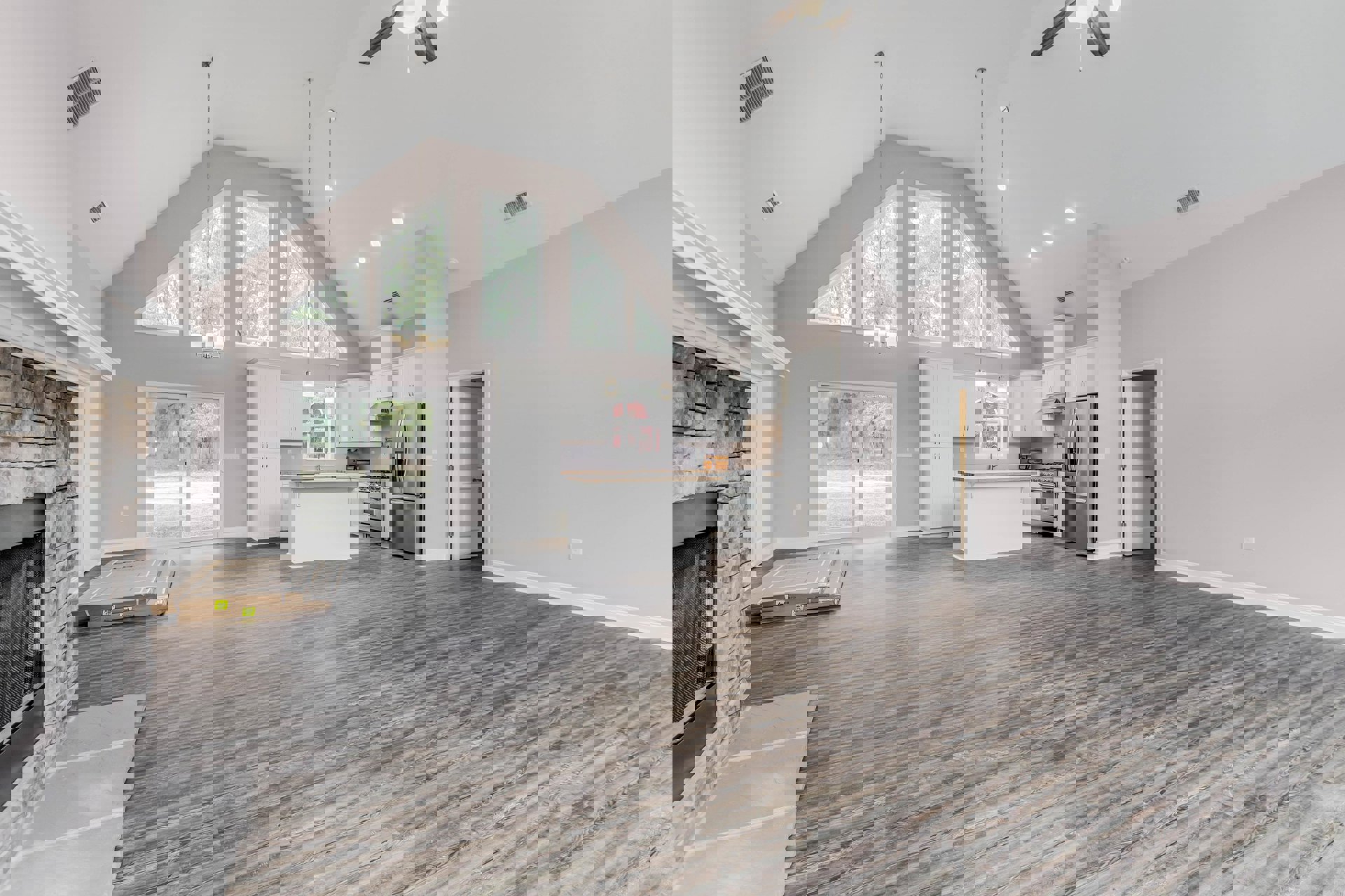 Spacious kitchen with wood flooring, central island, stainless steel refrigerator, ceiling vent, fireplace, and large window overlooking trees; cardboard moving boxes stacked near