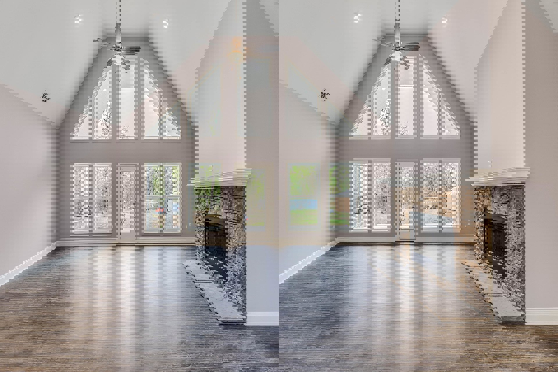 Living room with hardwood floors, stone fireplace featuring a black frame, ceiling fan with light fixture, and double doors opening to backyard view