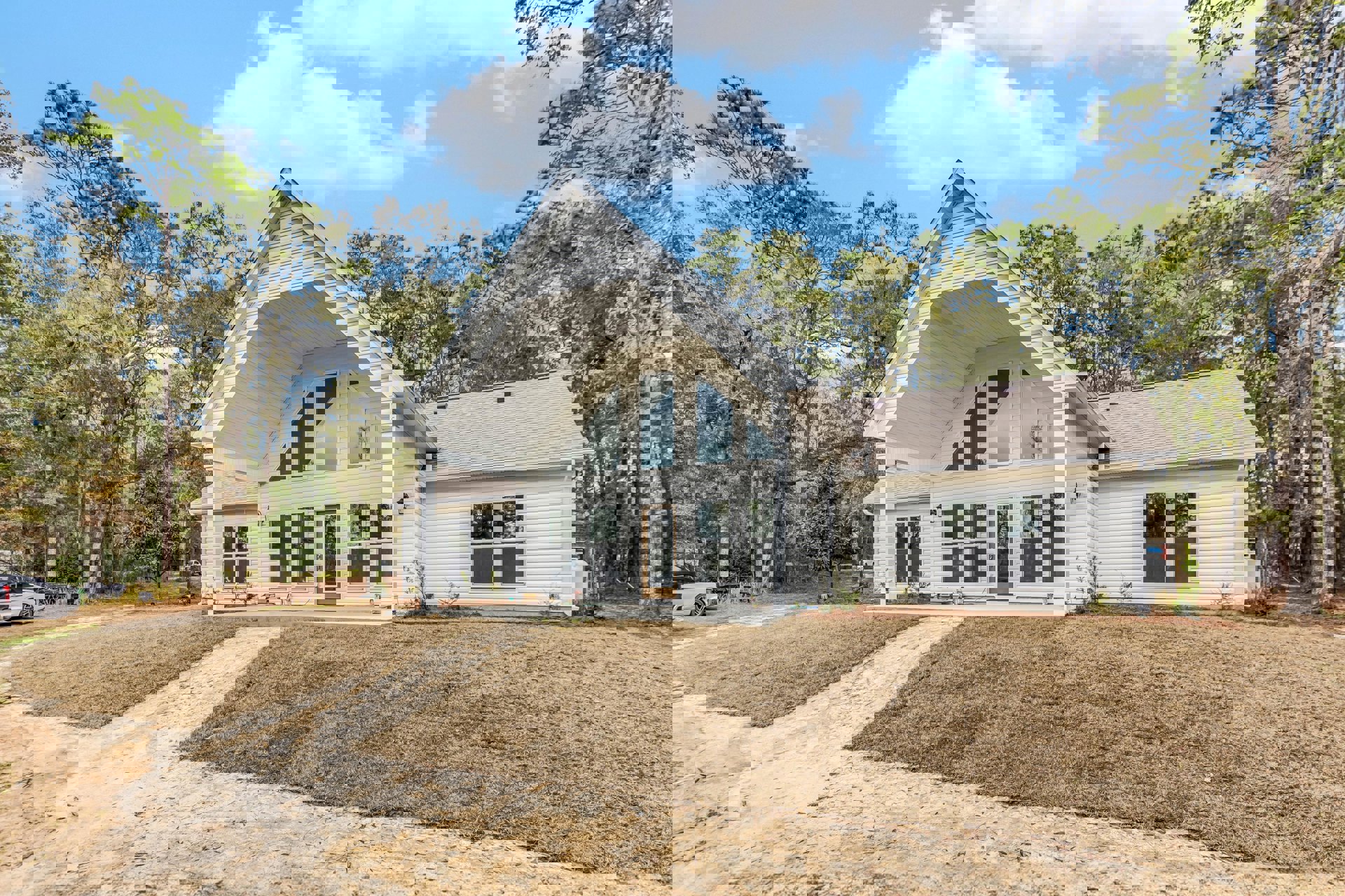 White house with triangular roof, black-topped car parked in driveway, grassy front yard, hill and mature trees in background, large windows facing outdoor landscape.