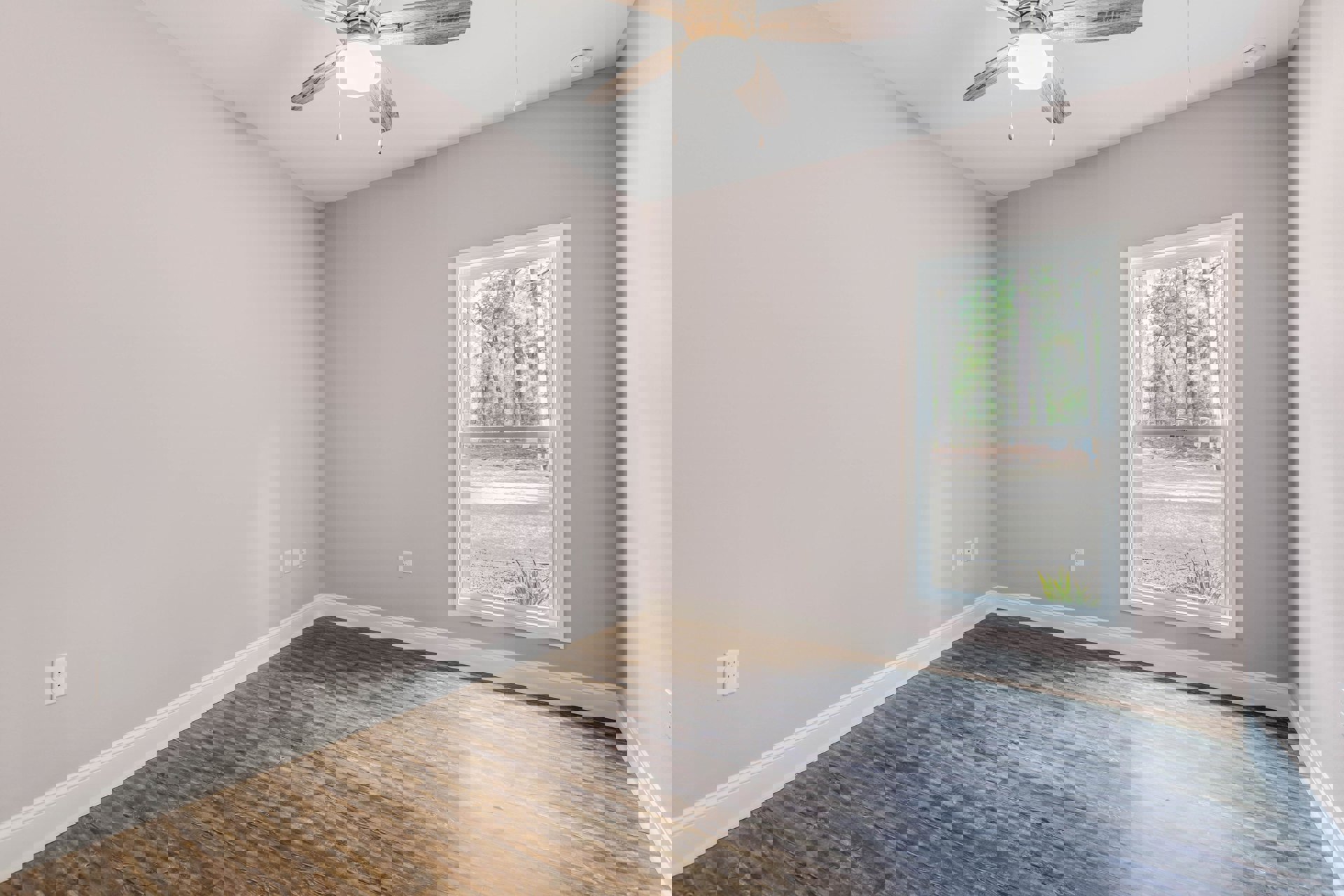 Wood flooring and white walls in a bright room with a ceiling fan and light fixture, large window overlooking trees, wall outlet visible near baseboard molding