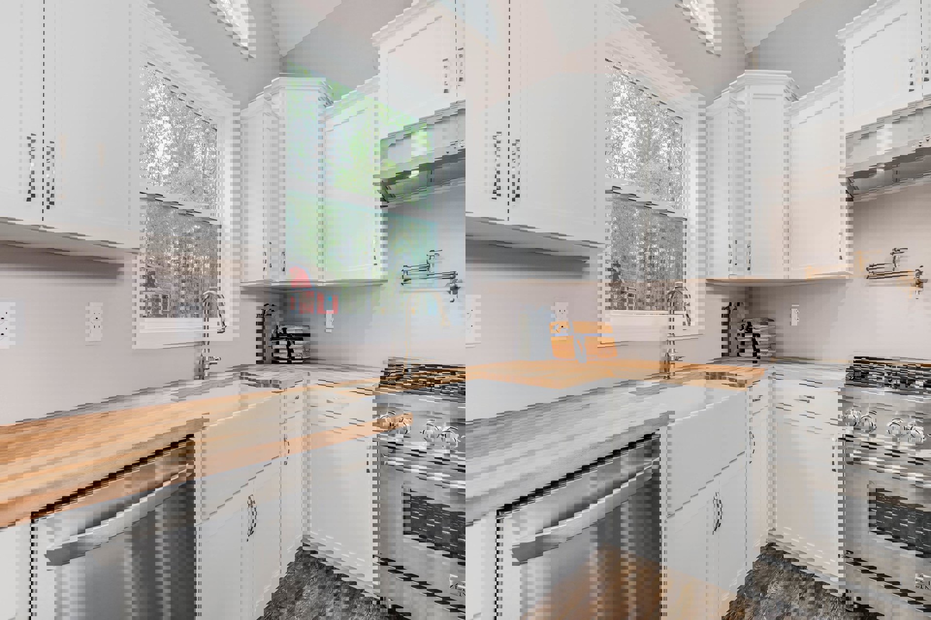 White kitchen cabinets with wooden countertops, stainless steel stove and dishwasher, white electrical outlet, window overlooking trees, wooden box with black ribbon on counter.