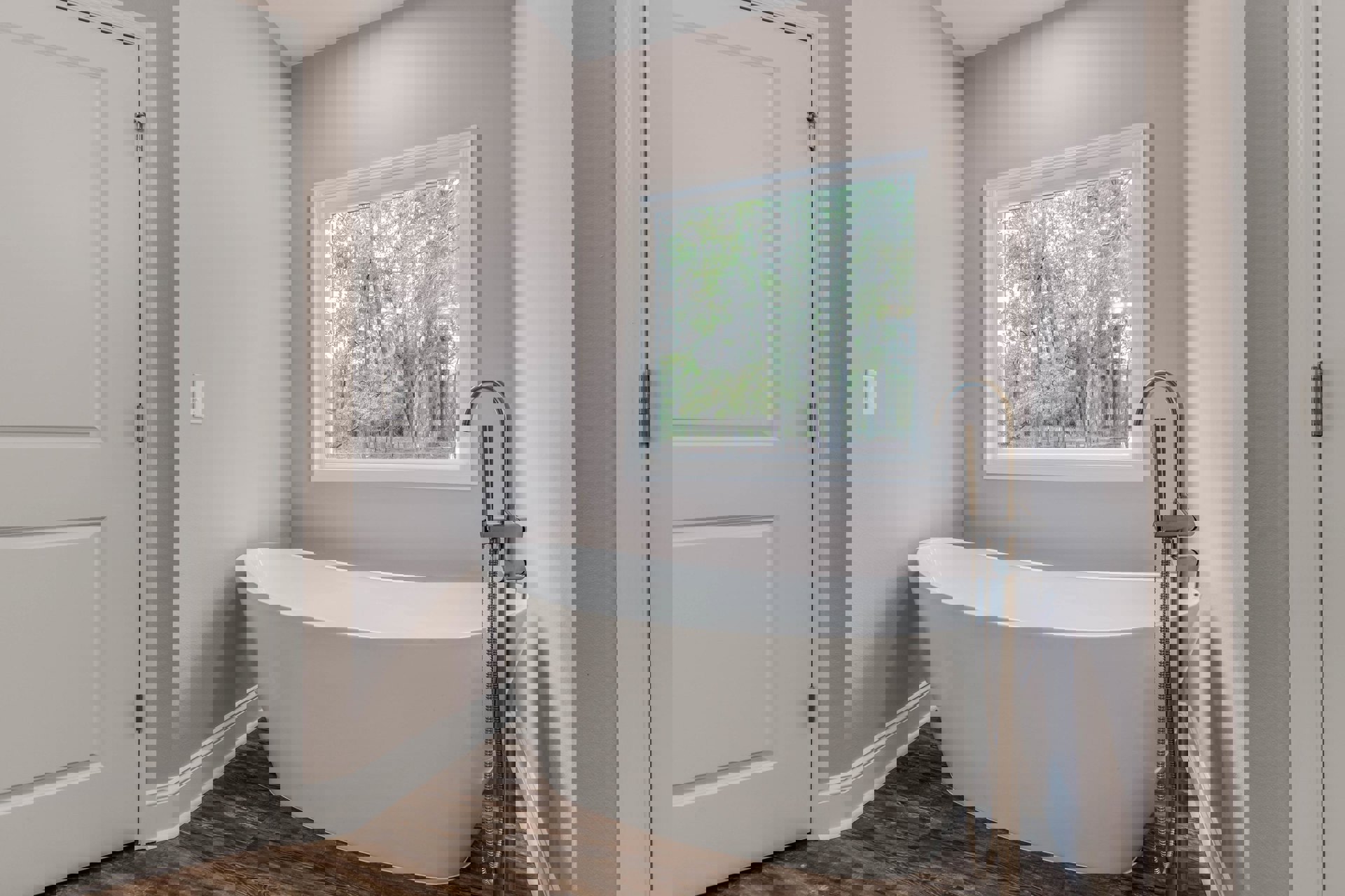 White freestanding bathtub with chrome faucet on tiled floor, large window overlooking trees, white paneled door visible in modern bathroom.