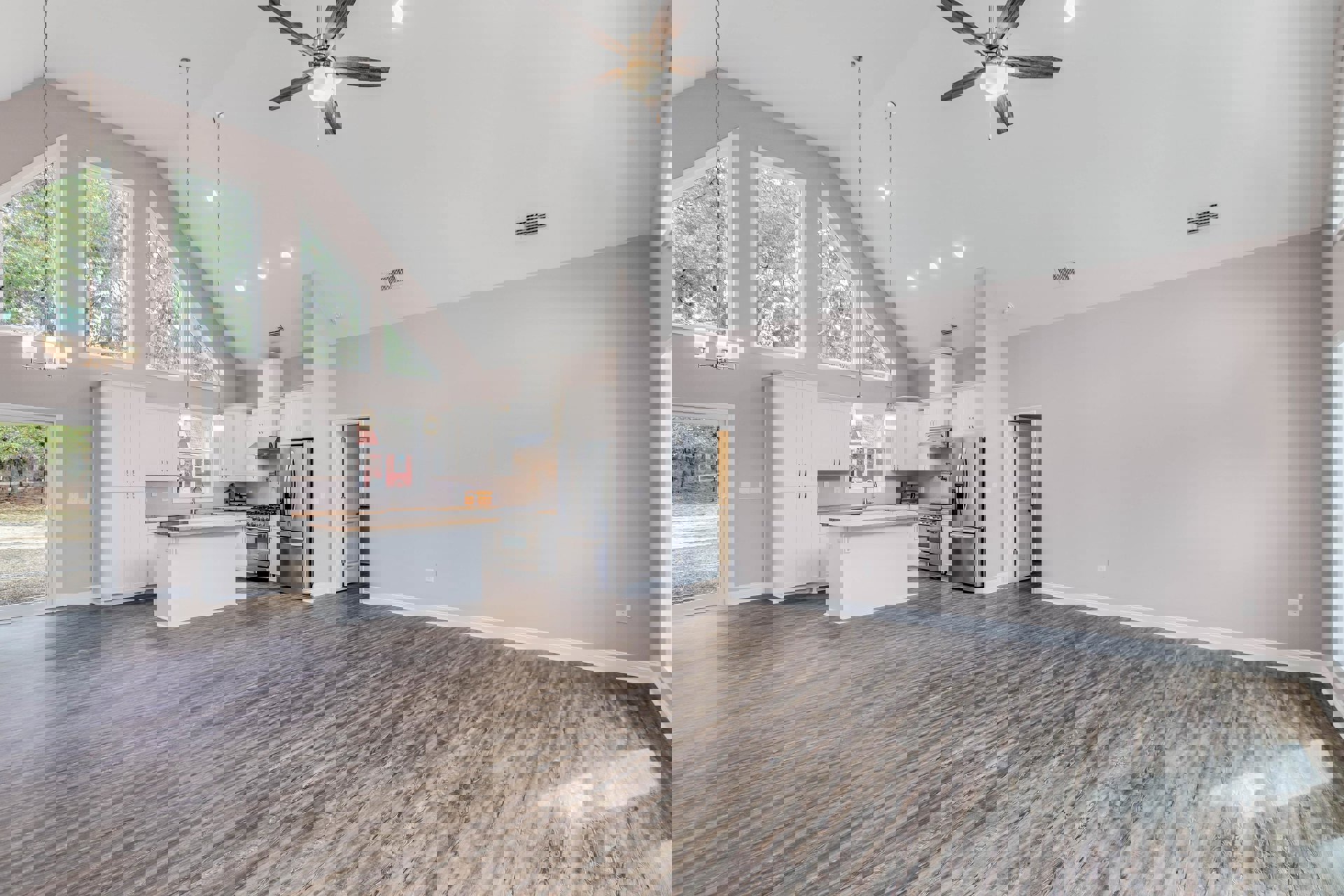 Spacious kitchen with wood flooring, white and wood kitchen island, stainless steel refrigerator with glass door, ceiling fan with light, large window showing trees outside