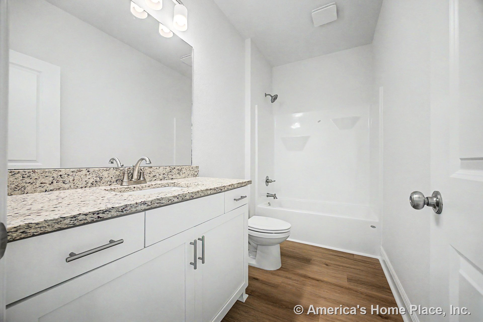 Granite vanity with chrome faucet atop white shaker cabinets, large wall mirror above, tub-shower combo with built-in shelves, wood-look flooring, modern overhead lighting, and