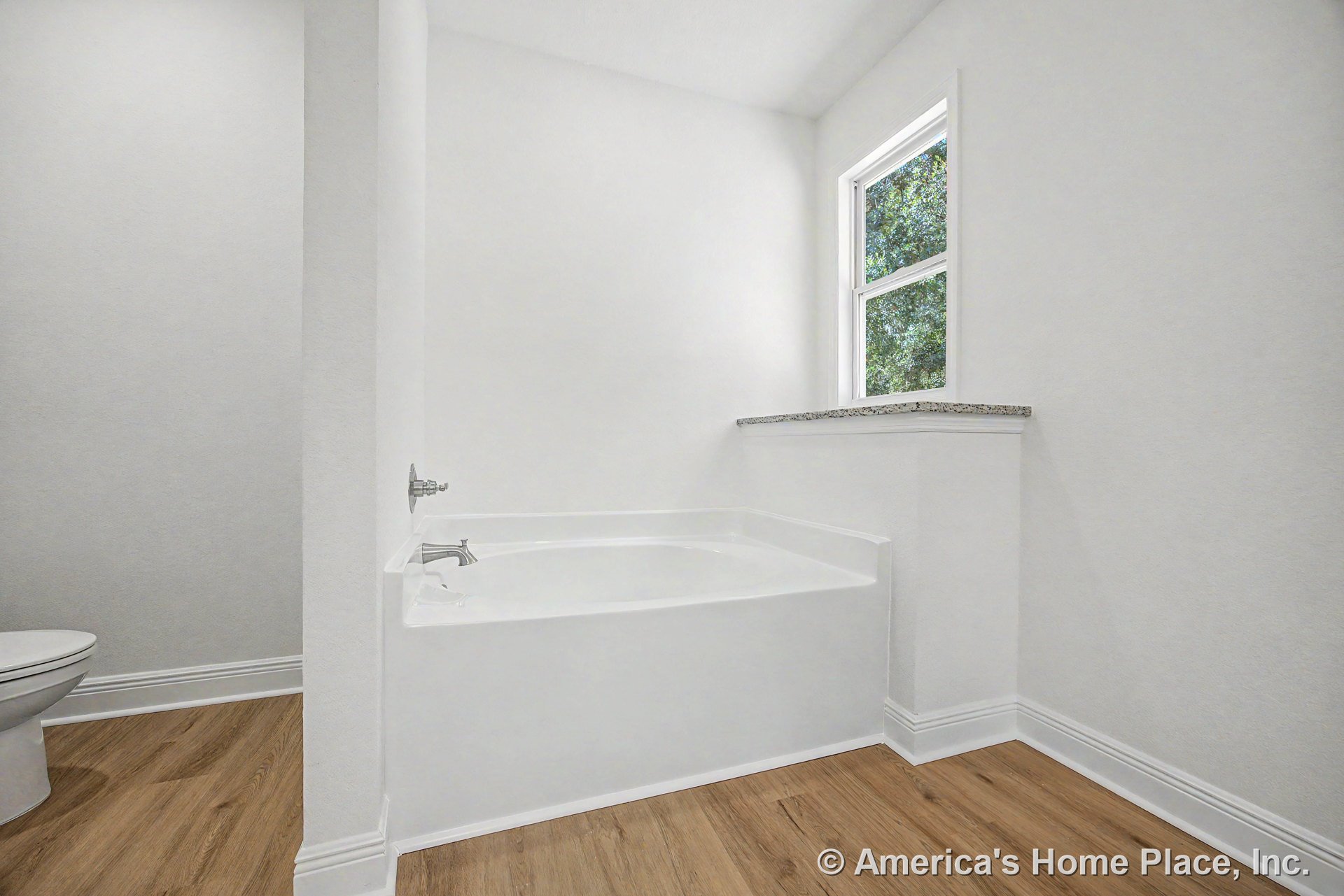 Built-in white soaking tub positioned beneath a double-hung window with a granite ledge, wood-look flooring, white baseboards, partial view of toilet, and white walls.