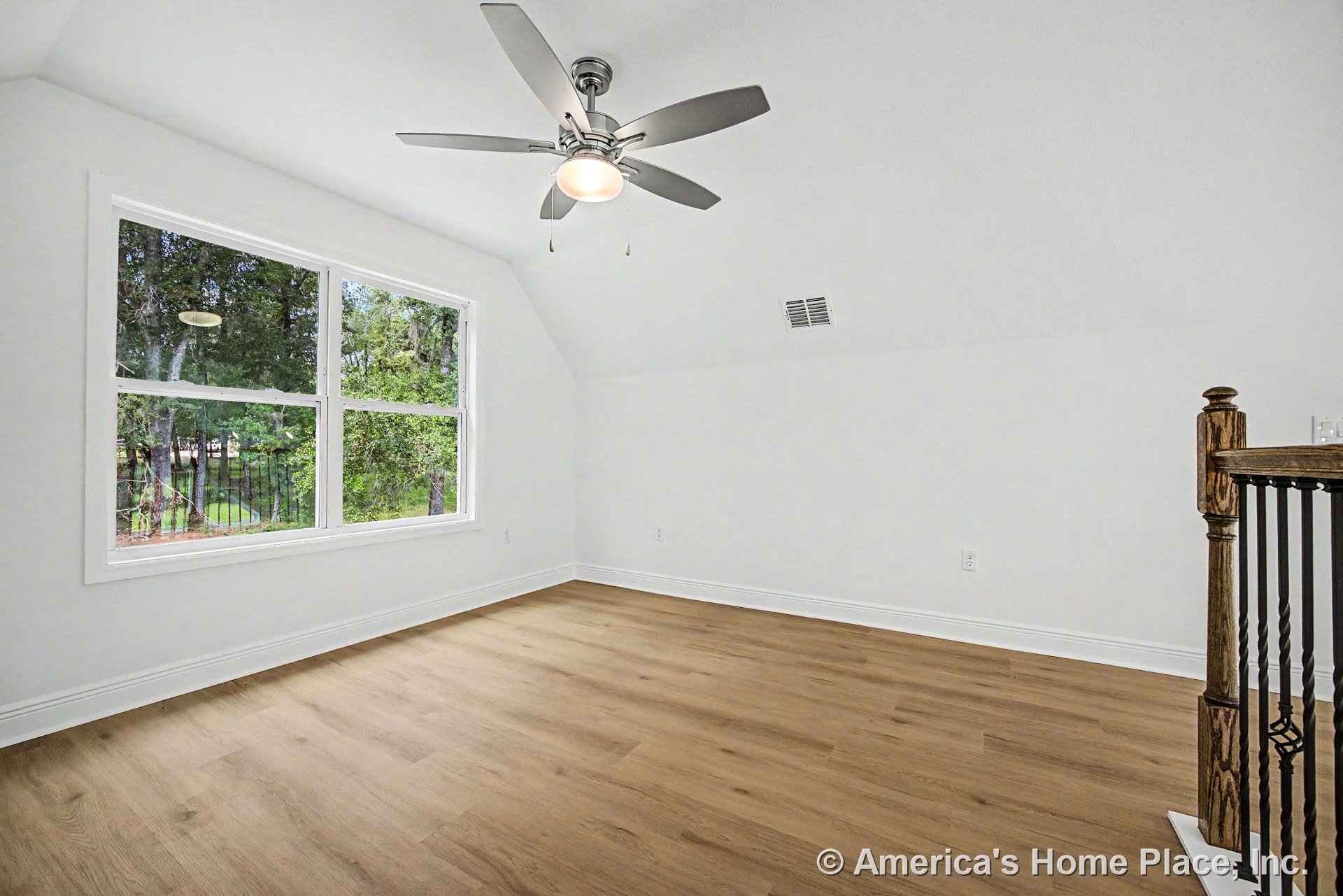 Upstairs room with large double window, light wood plank flooring, white baseboards, ceiling fan with light, angled ceiling, wood and iron stair railing, white walls, and vent