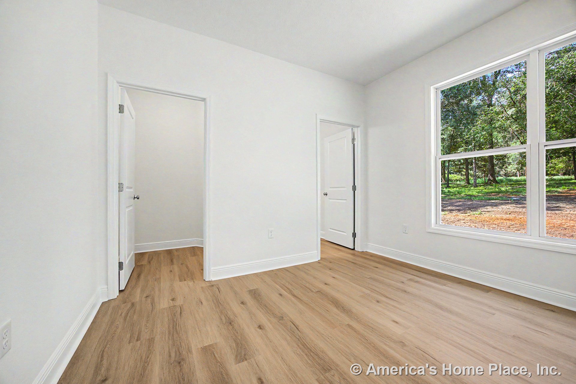 Bedroom with light wood-look flooring, large double window framed by white trim, smooth white walls, white baseboards, and two white panel interior doors.