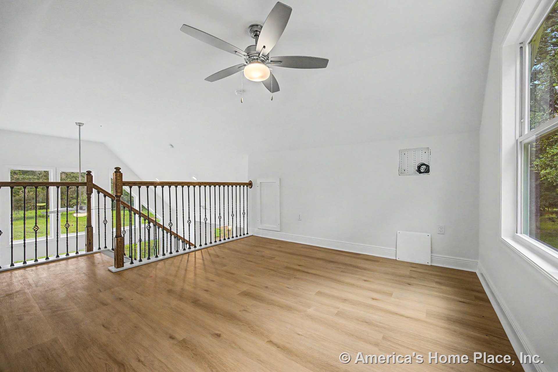 Open loft with light wood plank flooring, white walls, large window with trim, metal and wood stair railing, ceiling fan featuring built-in lighting, and abundant natural light.
