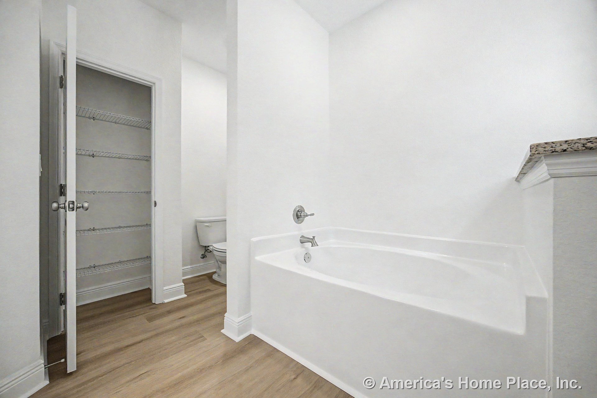 Built-in white soaking tub with chrome faucet beside granite countertop edge, adjacent to toilet and closet with wire shelving; wood-look flooring, white trim, and baseboards