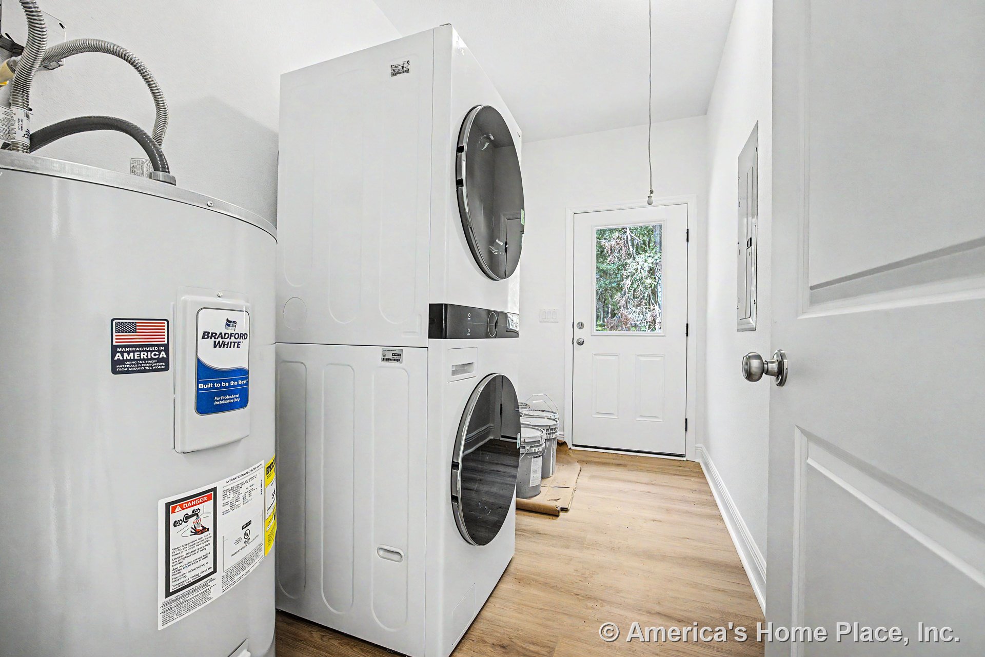 Stacked washer and dryer beside water heater tank, white exterior door with glass window, light wood flooring, white walls and trim, electrical panel mounted on wall.