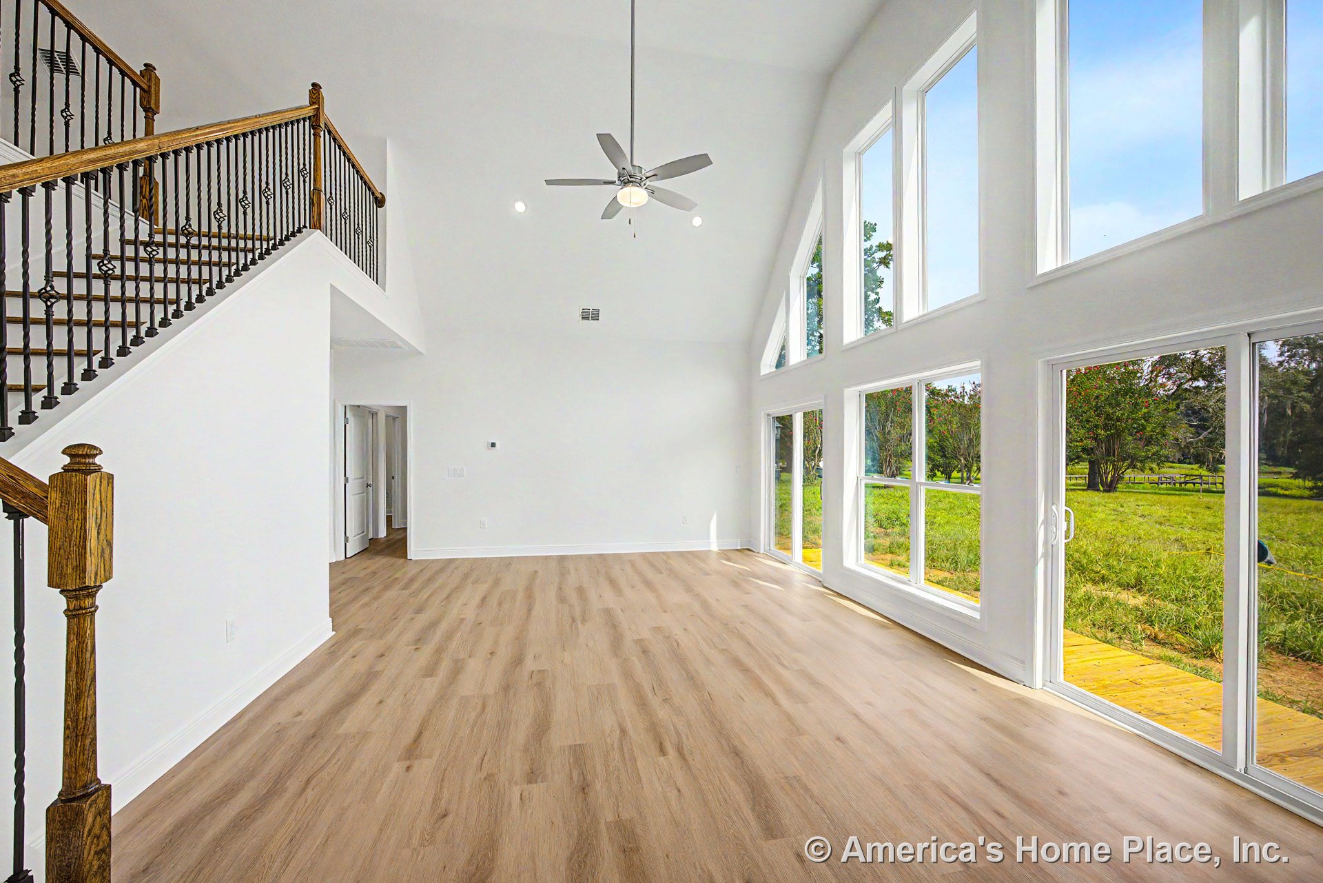 Vaulted ceiling with ceiling fan, expansive floor-to-ceiling windows, light wood plank flooring, white interior walls, iron and wood staircase railing, sliding glass door leading
