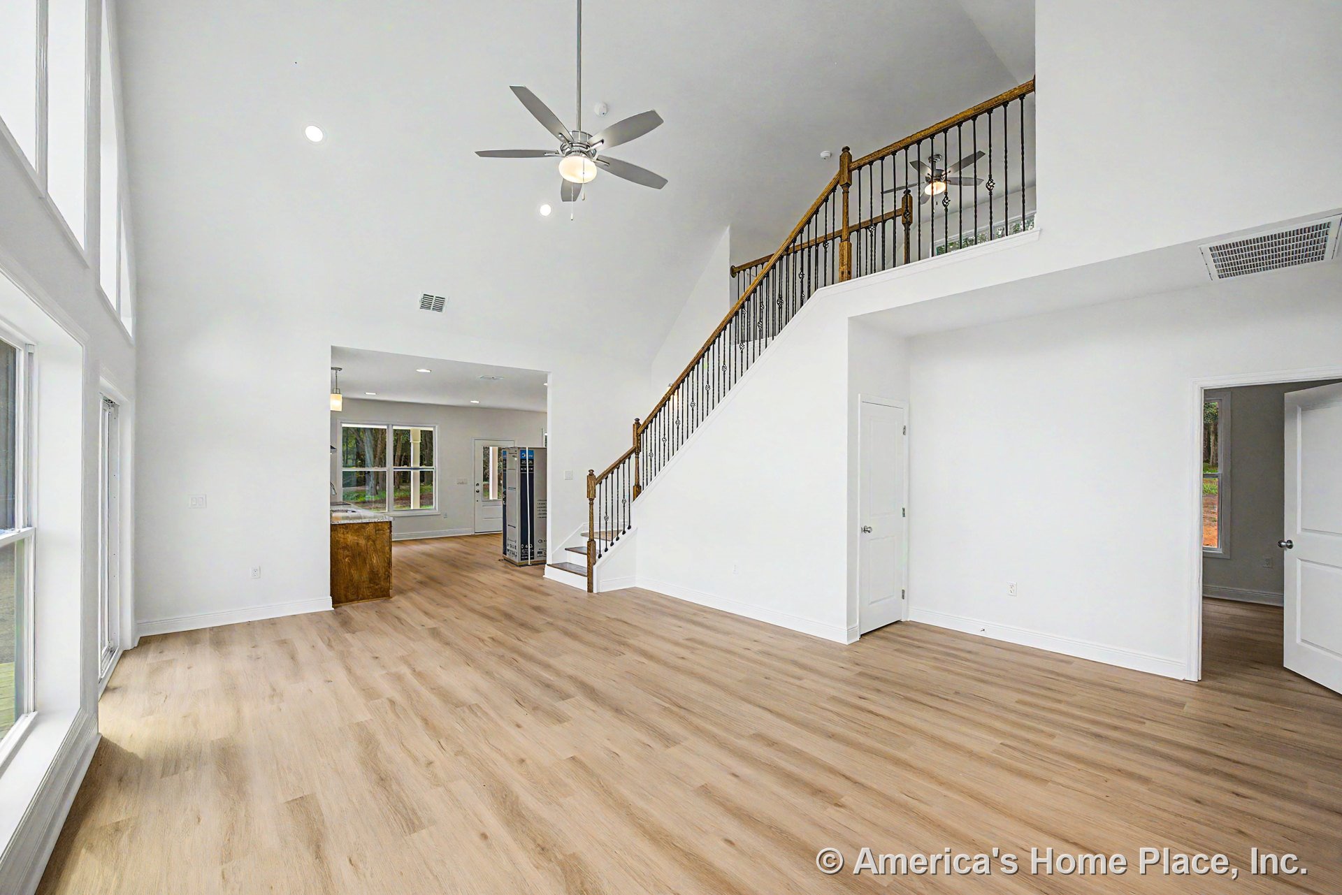 Open living room with light wood flooring, vaulted ceiling and ceiling fan, expansive window wall, staircase featuring iron balusters, white interior trim and baseboards, interior