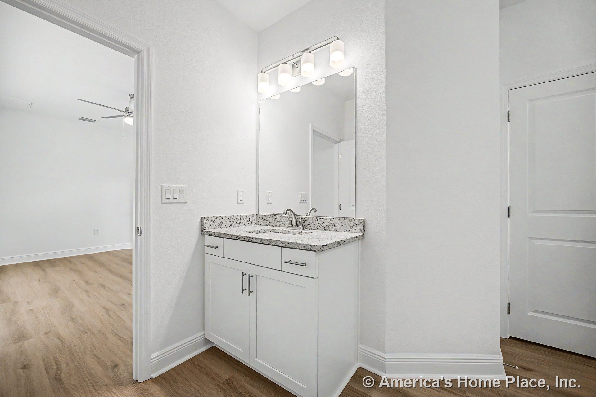 White shaker-style vanity with granite countertop, large wall-mounted mirror, four-light vanity fixture, wood-look flooring, white trim and doors.