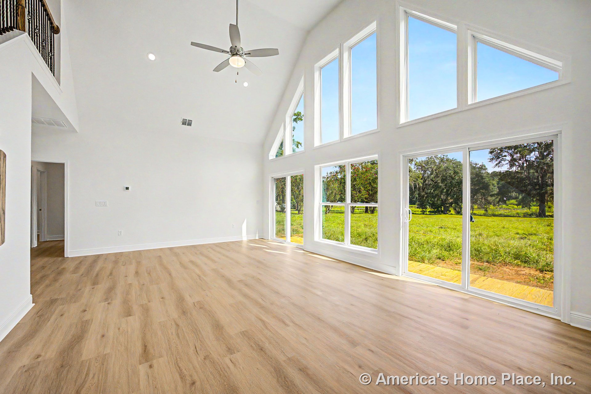 Vaulted ceiling living room with large triangular floor-to-ceiling windows, sliding glass doors, light wood flooring, white trim, ceiling fan with light, and abundant natural