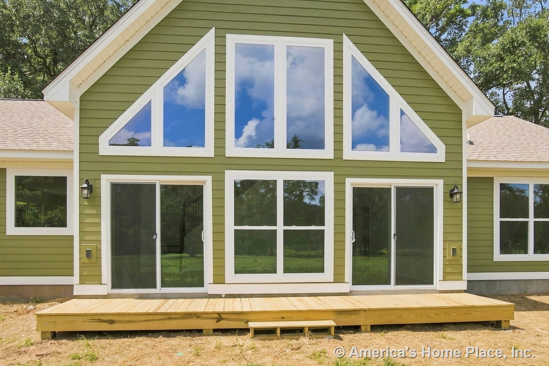 Green horizontal siding with white trim accents, large triangular and rectangular geometric windows, sliding glass doors leading to a wooden deck with steps, sloped roofline, and