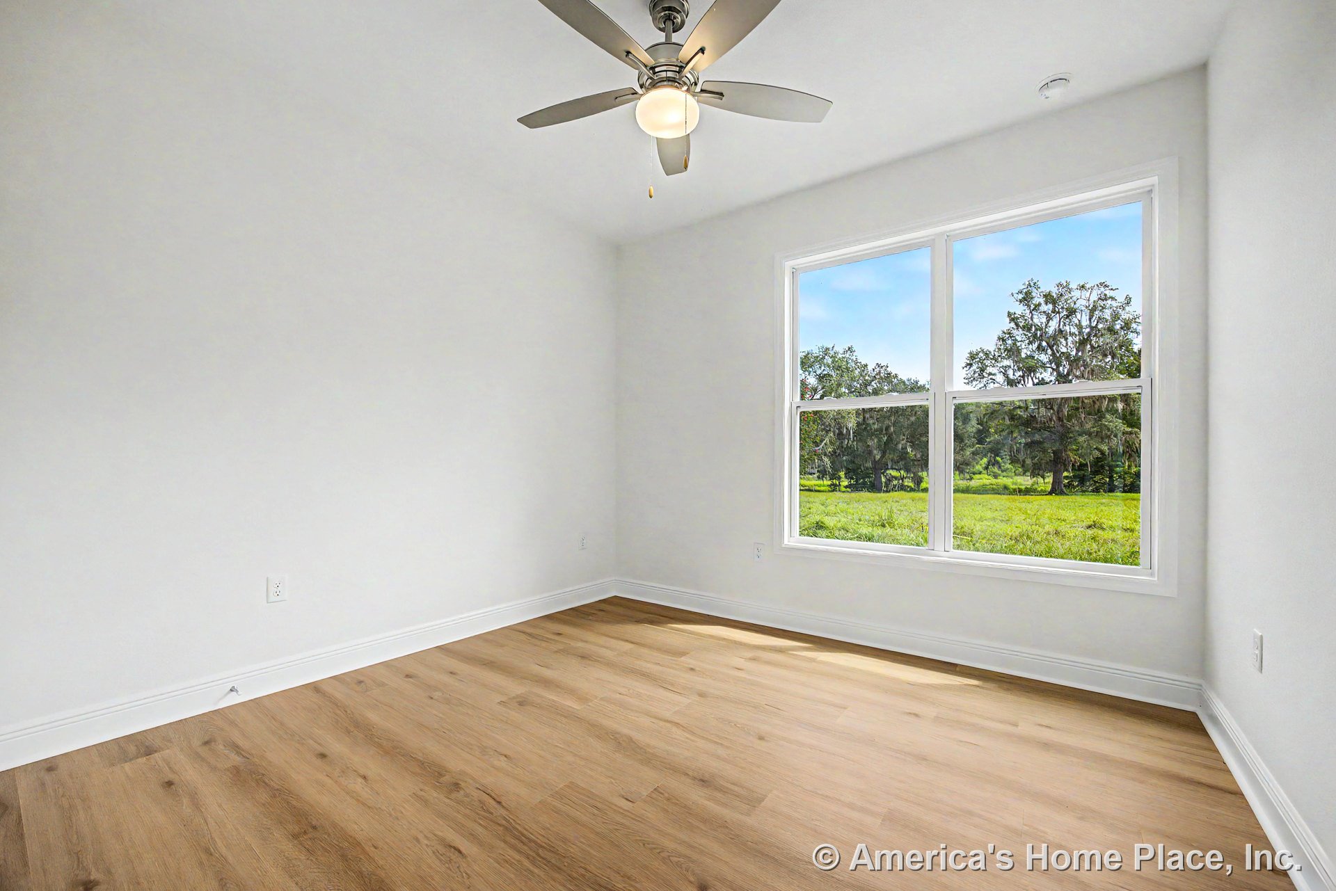 Bedroom with light wood plank flooring, large double window framed by white baseboards and trim, ceiling fan with integrated lighting, smooth white walls, and abundant natural