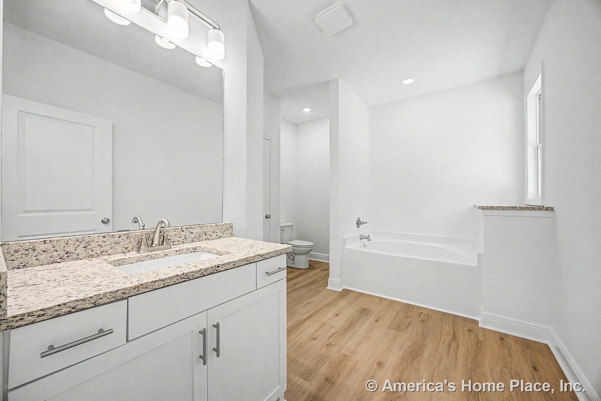 Granite vanity countertop with single sink and chrome faucet, white shaker cabinets, built-in bathtub, wood-look flooring, large wall mirror, recessed ceiling lights, window with
