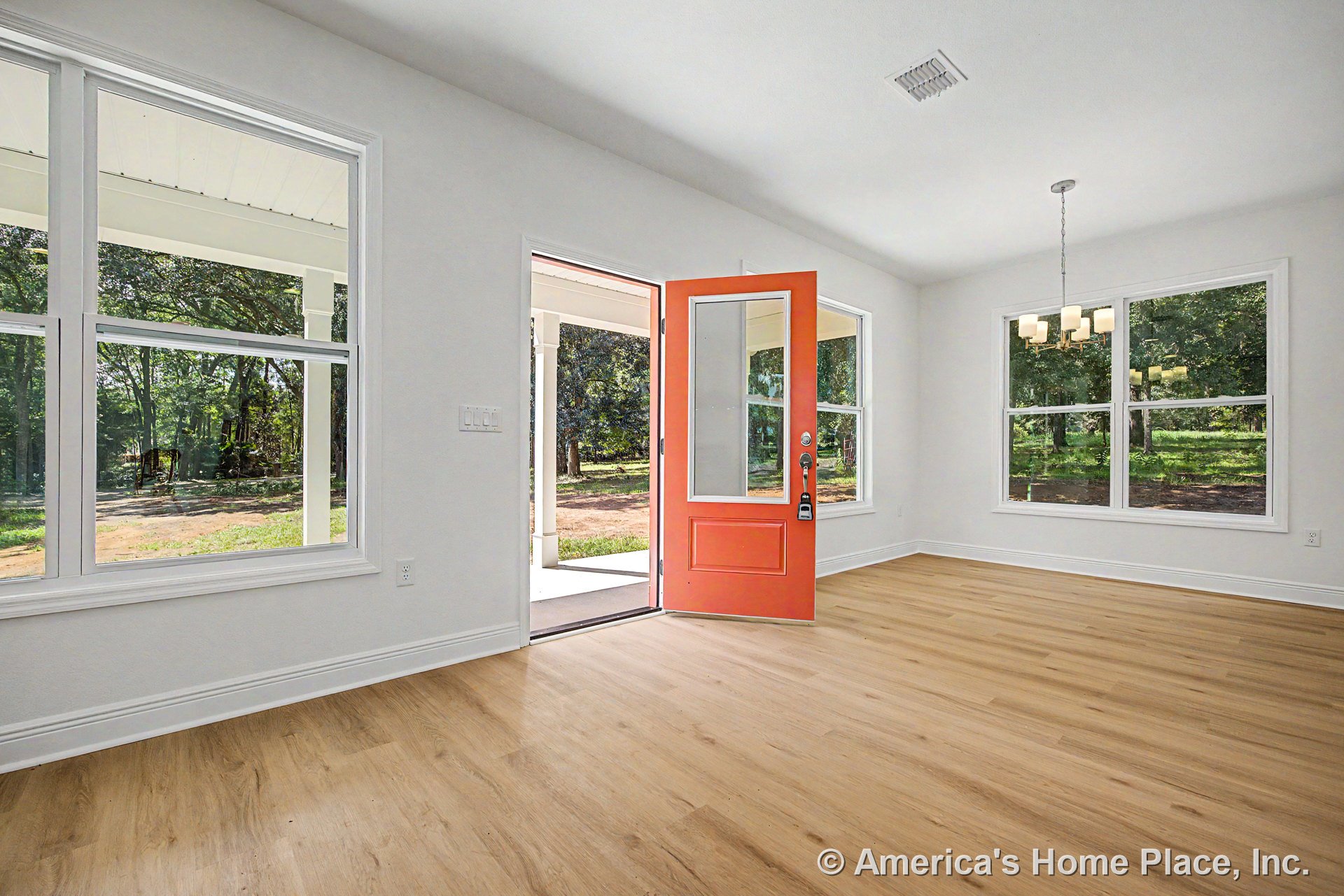 Red front door with white trim opens to a foyer featuring large double-pane windows, light wood plank flooring, modern chandelier, white baseboards, and covered porch visible