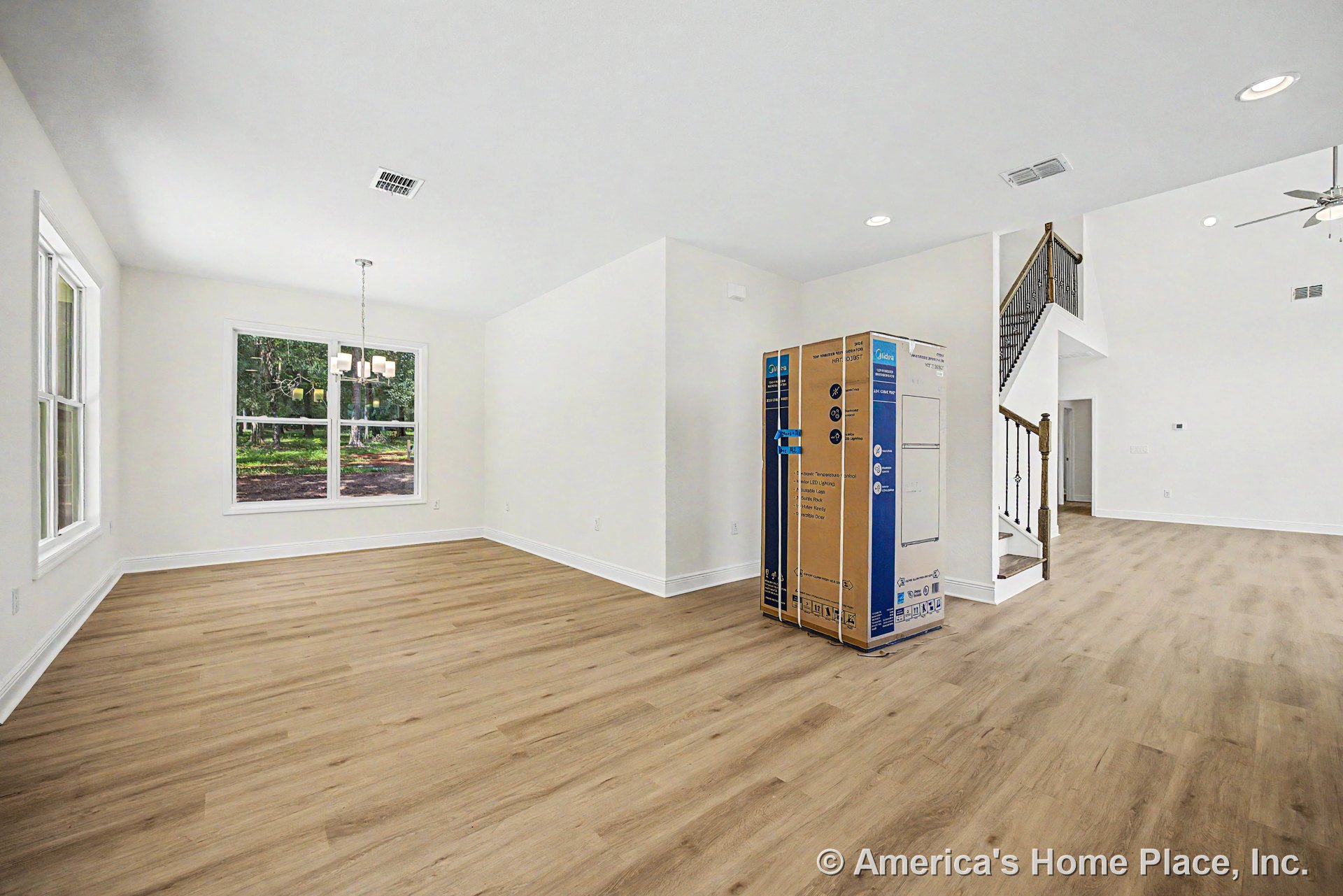 Open living area with light wood plank flooring, large double windows with white trim, recessed ceiling lights, staircase featuring wood steps and iron balusters, dining area with