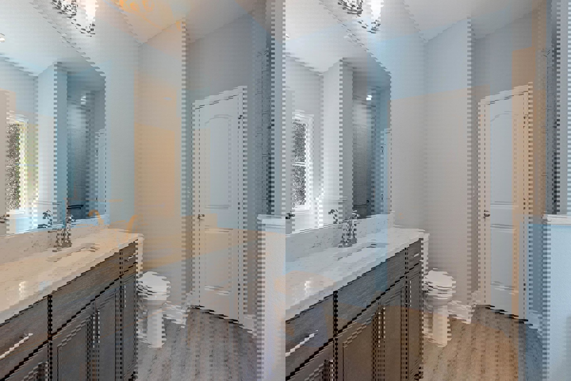 Bathroom with wood flooring, white baseboards, white toilet, sink with cabinetry, silver faucet, large mirror, white door with silver knob, window showing trees outside