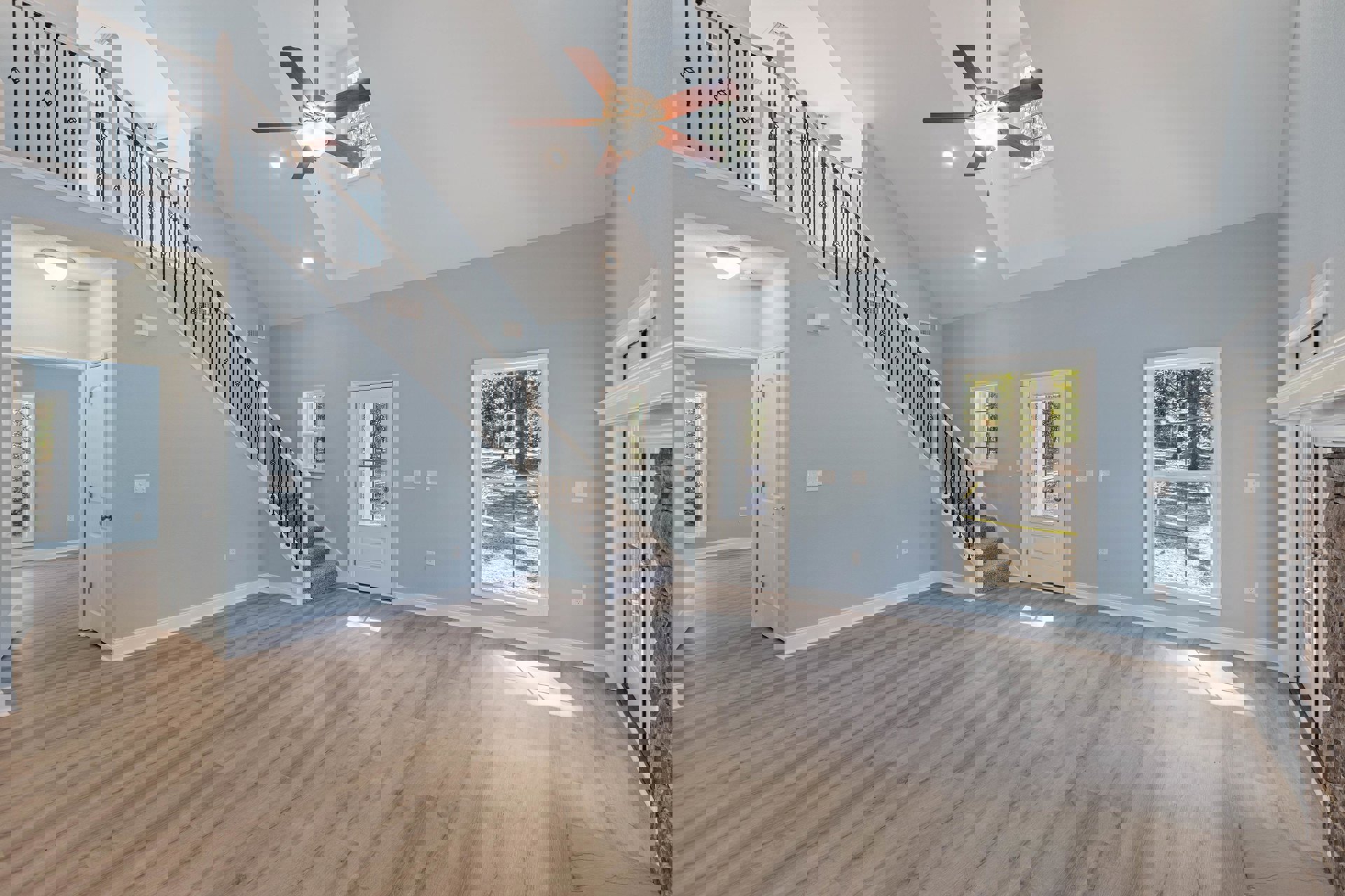 Hardwood floor living area featuring a ceiling fan with light fixture, white door with glass window, staircase with wooden steps, and large window overlooking forest scenery