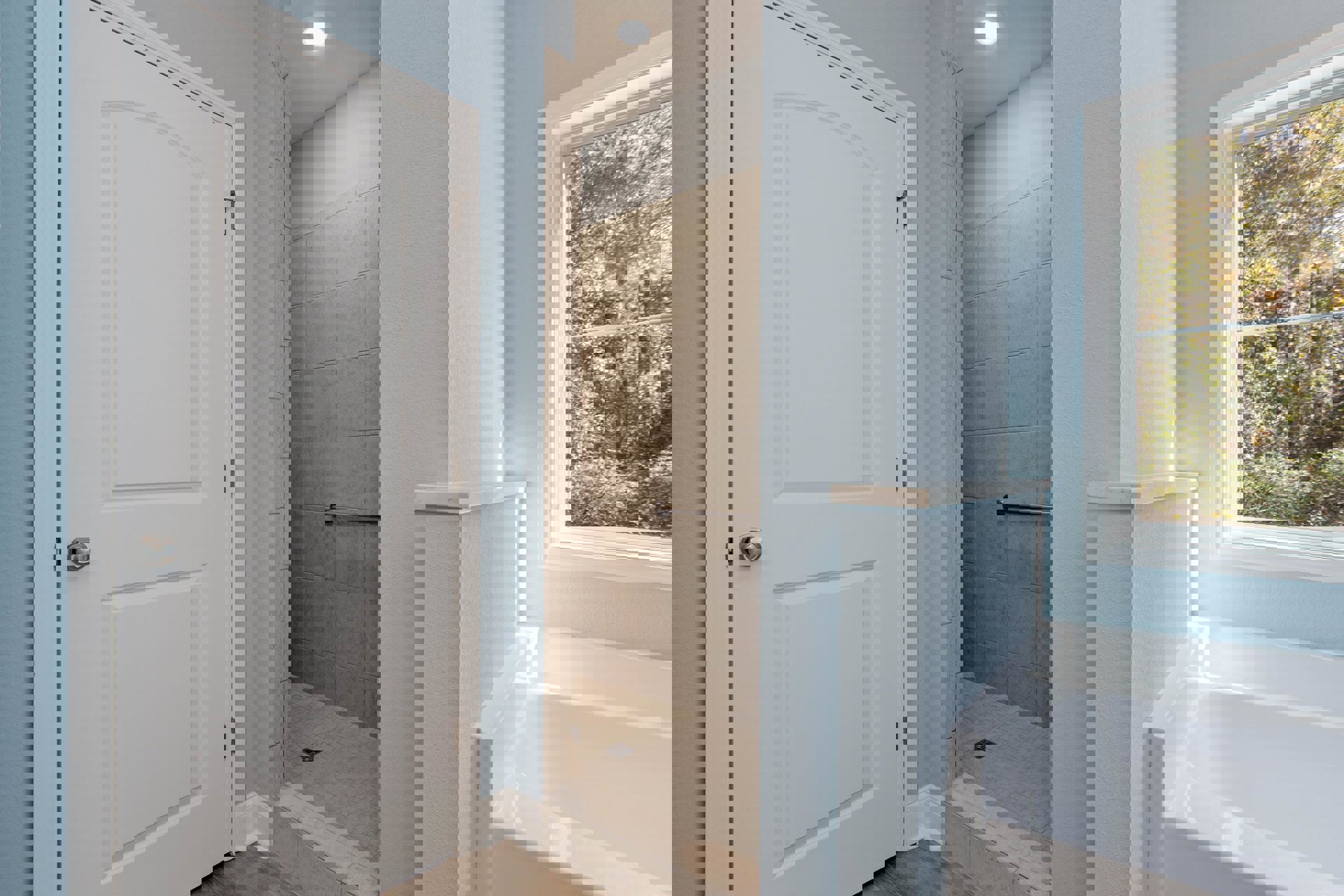 Bathroom with white bathtub and glass shower, large window showing trees outside, white paneled door with silver handle, ceiling light, light-colored tile walls and floor