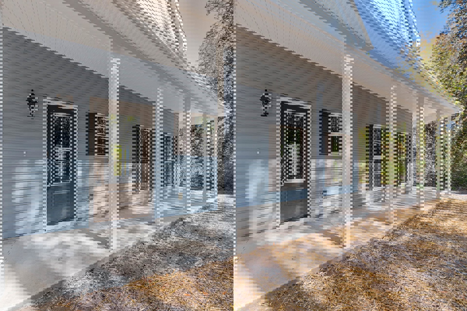 White house exterior with covered porch, white columns, glass-paneled front door, white-framed window, brown leaves scattered on ground, and outdoor lamp.