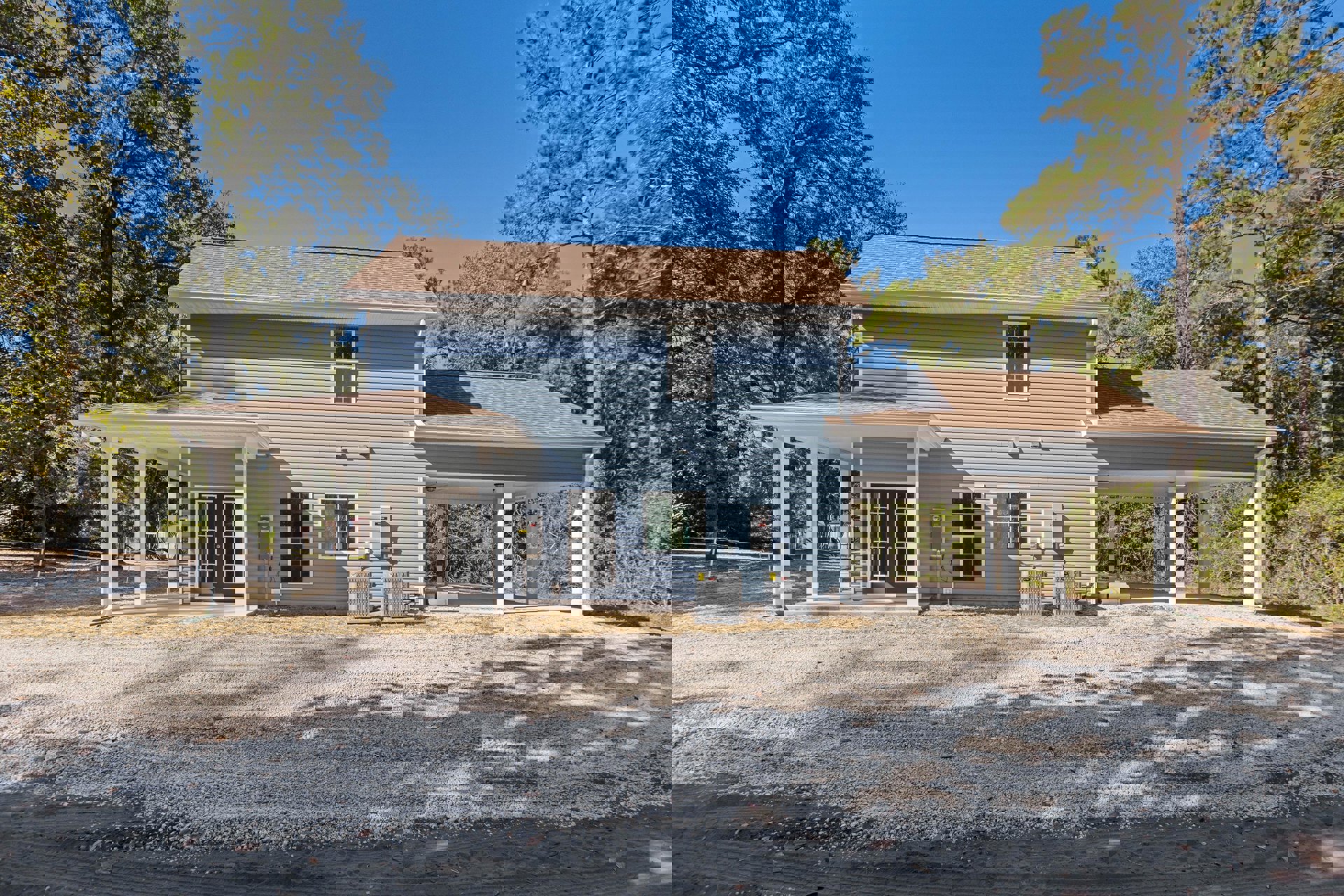 Modern home exterior with light-colored siding, covered patio supported by columns, white-framed windows with grid detailing, gravel landscaping, and visible electrical boxes near