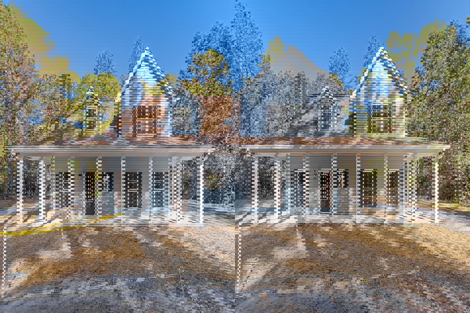 Blue house with white-trimmed windows, covered porch, and driveway bordered by straw and grass, fenced area in foreground, Sidney Lanier Cottage visible in background.