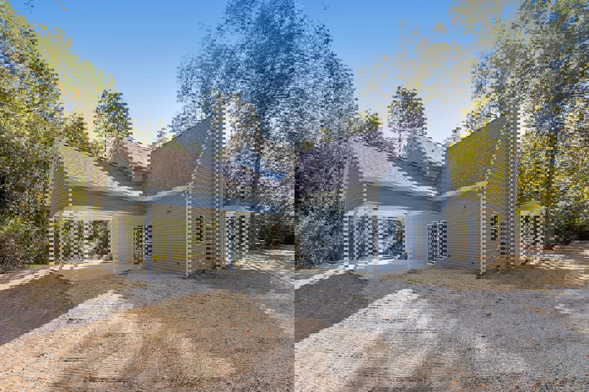 Covered patio with white pillars, gravel driveway, mature trees, and cottage-style home exterior