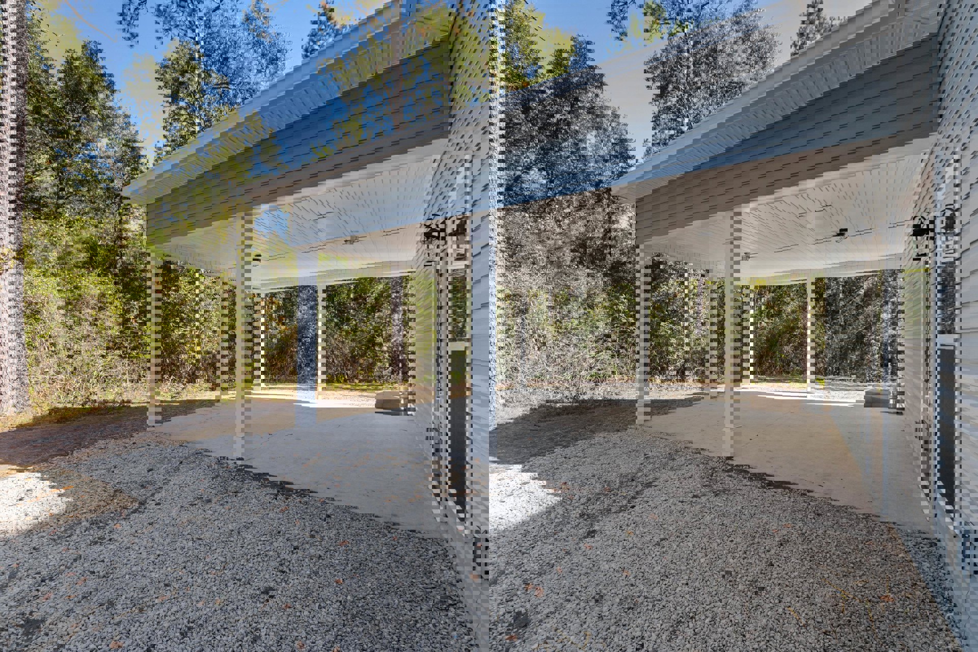 Concrete patio with white roof and pillars, shaded by trees in the background, outdoor lamp mounted near window, blue sky visible above.