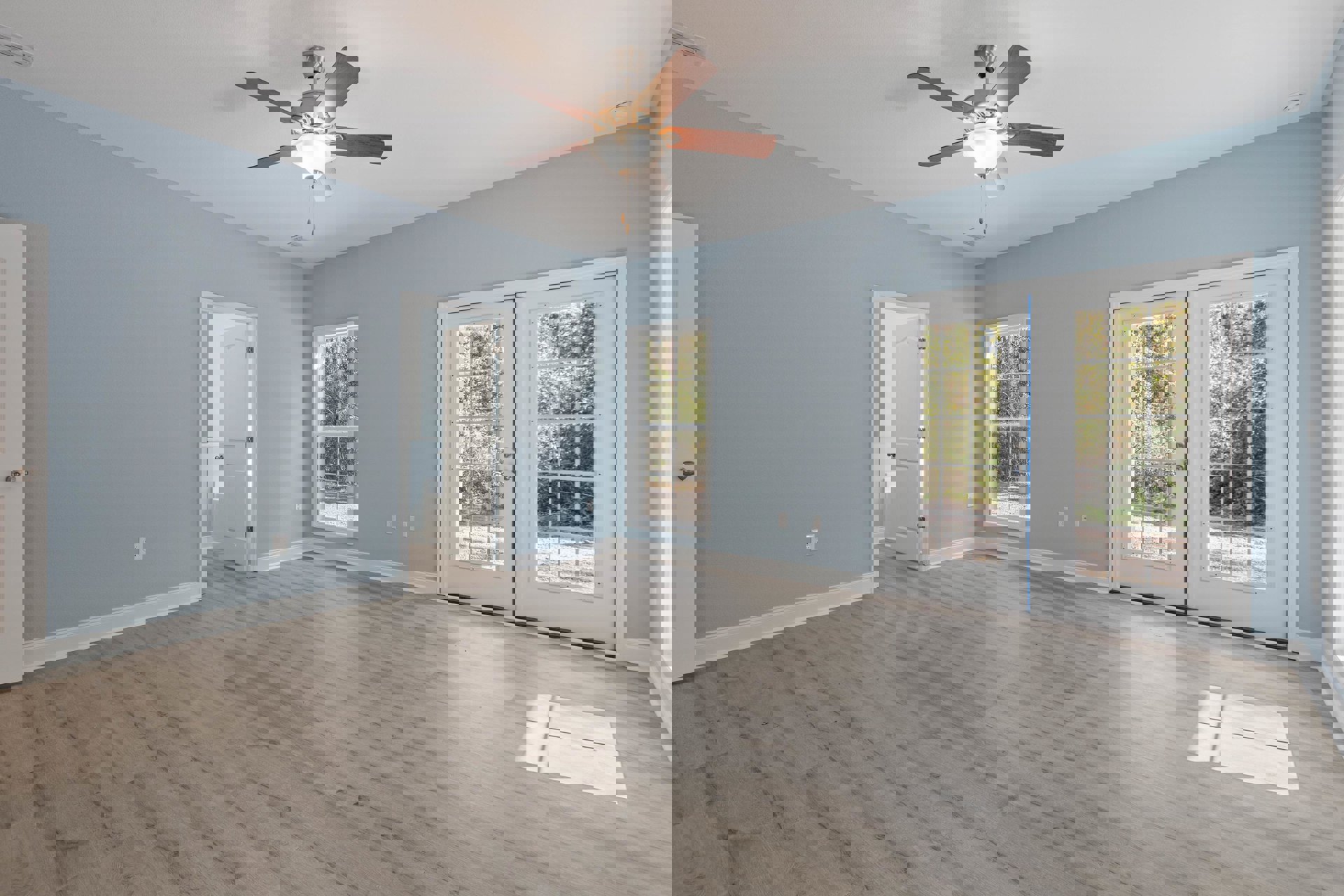 Ceiling fan with light fixture above wood flooring, double glass-paneled doors, white walls, window showing trees outside, silver door knob