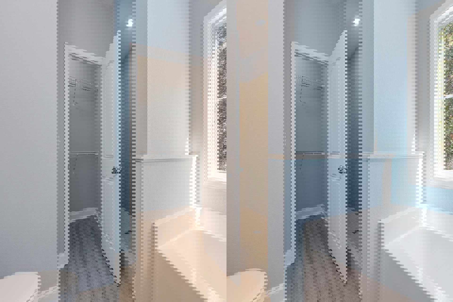 Bathroom with white tile walls, open door, white bathtub in sunlight, window with white frame, close-up of toilet seat, adjacent closet with white door, white clothes rack visible