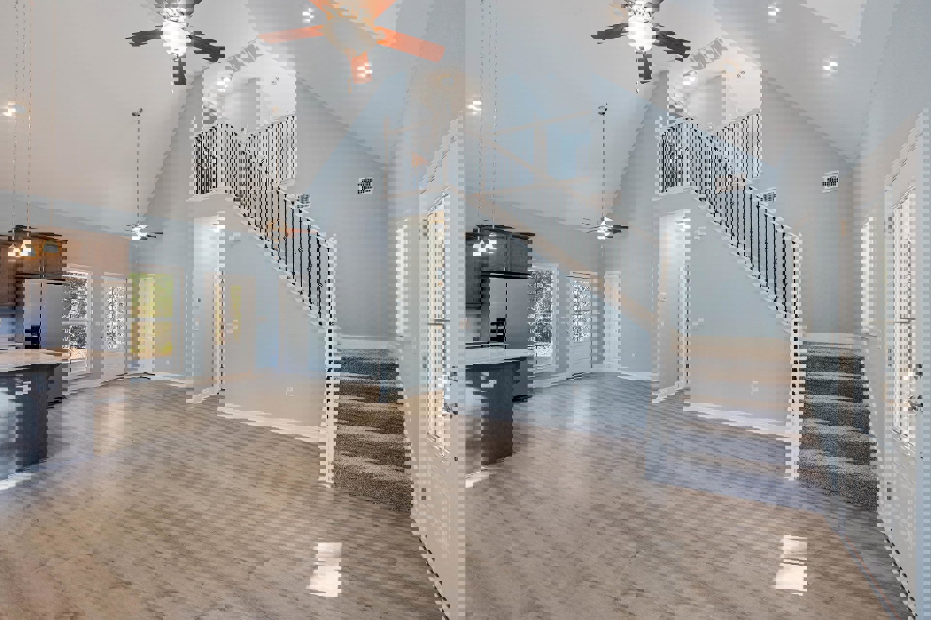 Open-concept living area with wood flooring, central staircase, ceiling fan with light fixture, kitchen island, white door with window, and large windows overlooking trees.