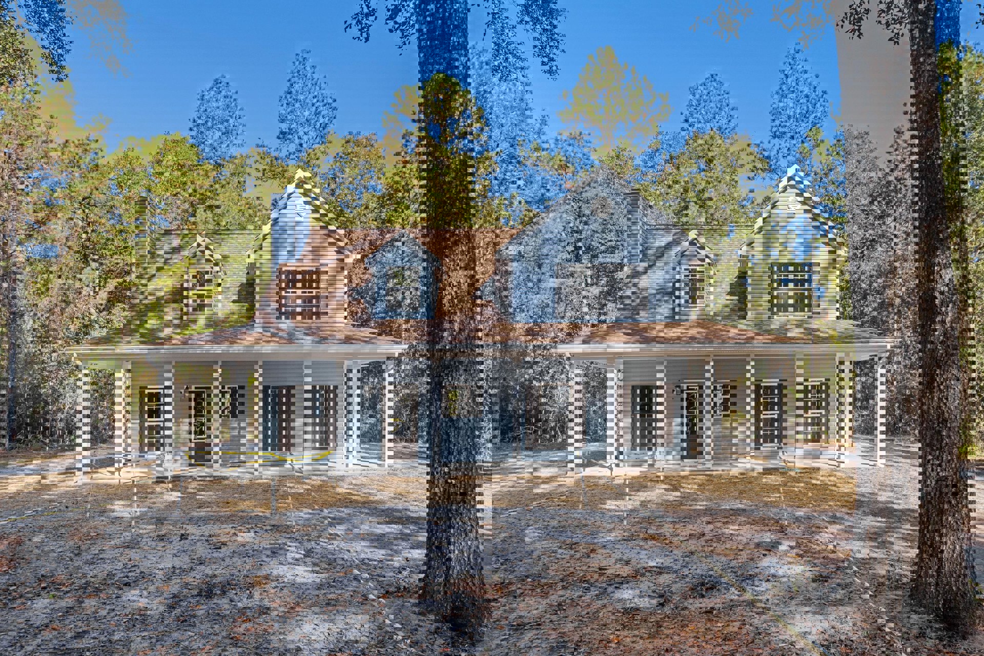 Blue siding house with brown roof, white-framed windows, yellow tape on one window, surrounded by mature trees, dirt yard in front.