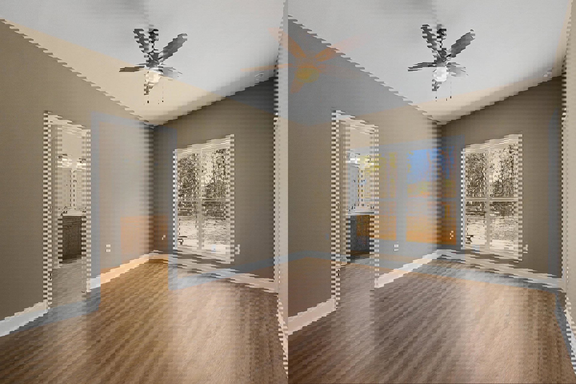 Ceiling fan with light fixture, wood flooring with white trim, large window overlooking trees, bathroom with double sink and two mirrors, partial view of interior fence