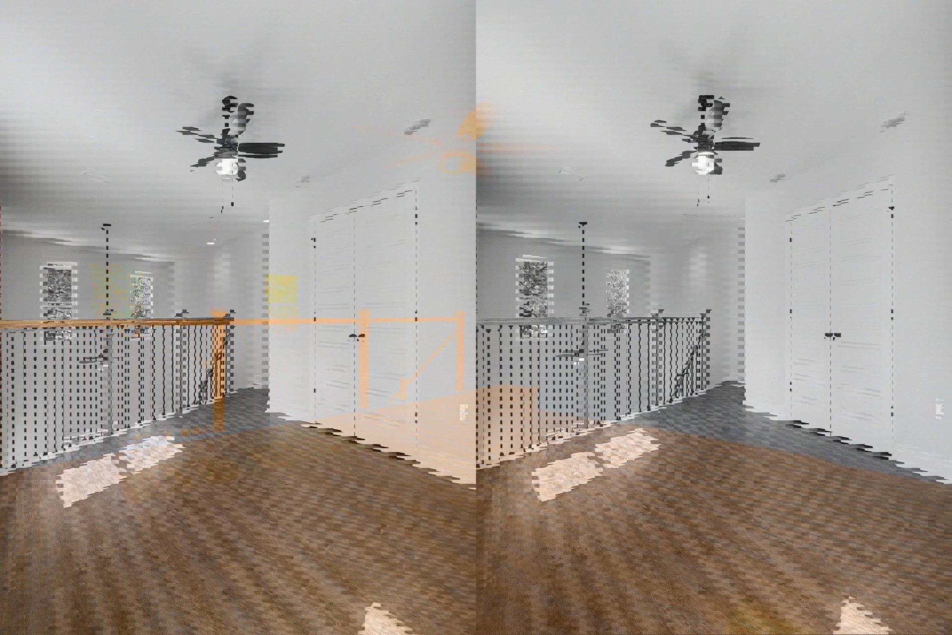 Wood floor with metal railing, ceiling fan with light fixture, white door with black knobs, window showing trees outside, plaster walls.