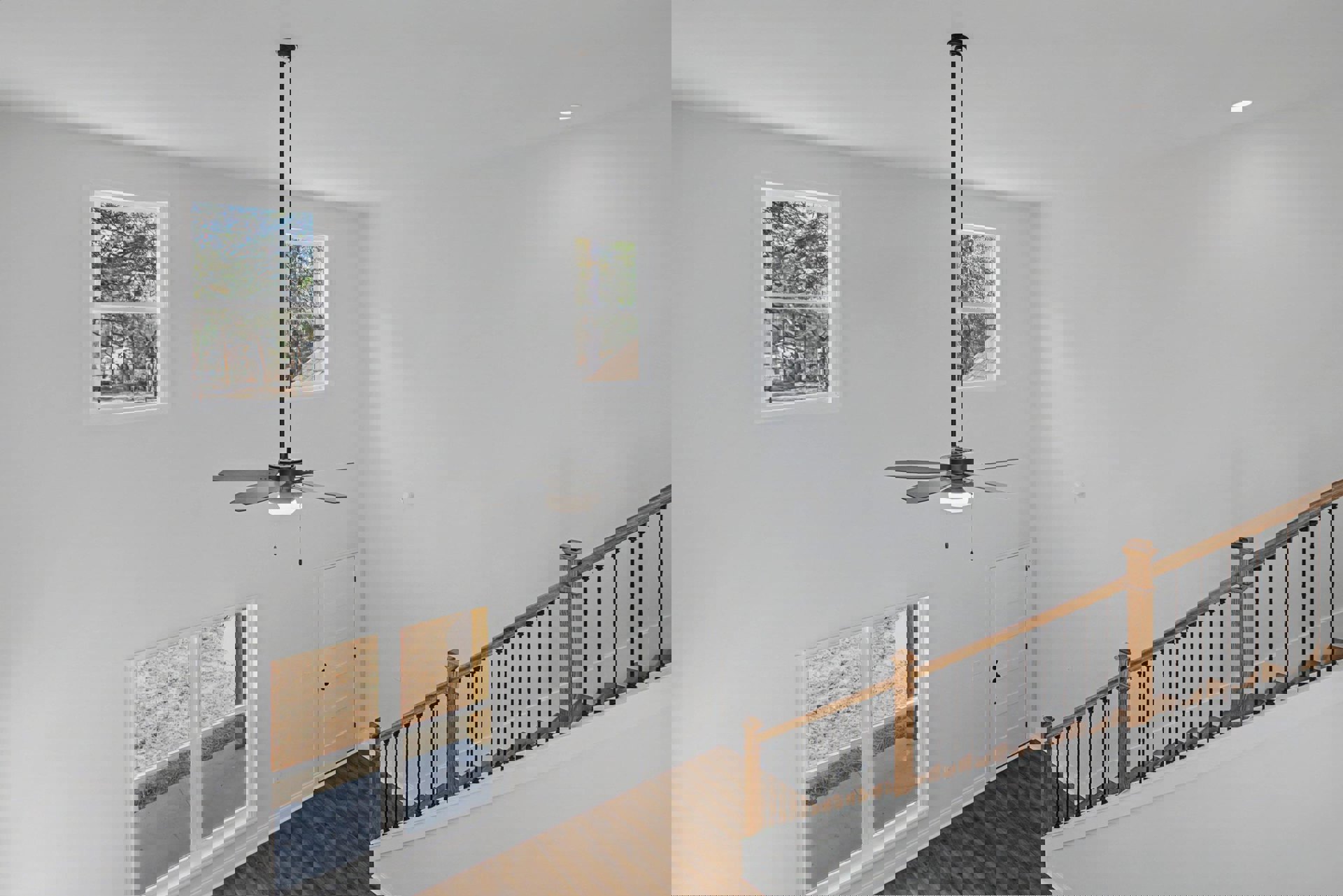 Ceiling fan mounted above a living area with wood-railed staircase, multiple windows showing views of trees and neighboring house, white door, plaster walls, and decorative